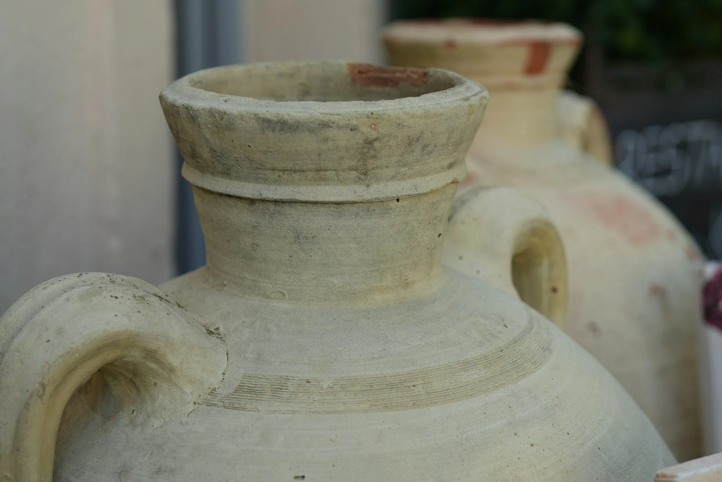Close-up of a large, ancient ceramic vessel with a rounded body, a narrow neck, and decorated with rings, displayed outdoors. It shows signs of aging with some chips and dirt, and there are other similar vessels in the background.