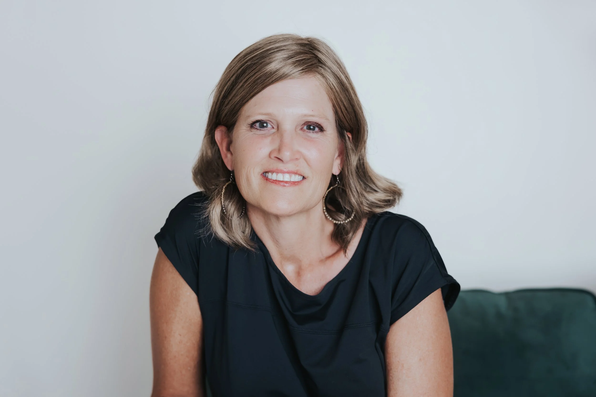 Headshot of a middle-aged woman with shoulder-length light brown hair, wearing a black top and earrings, smiling at the camera against a plain light background.