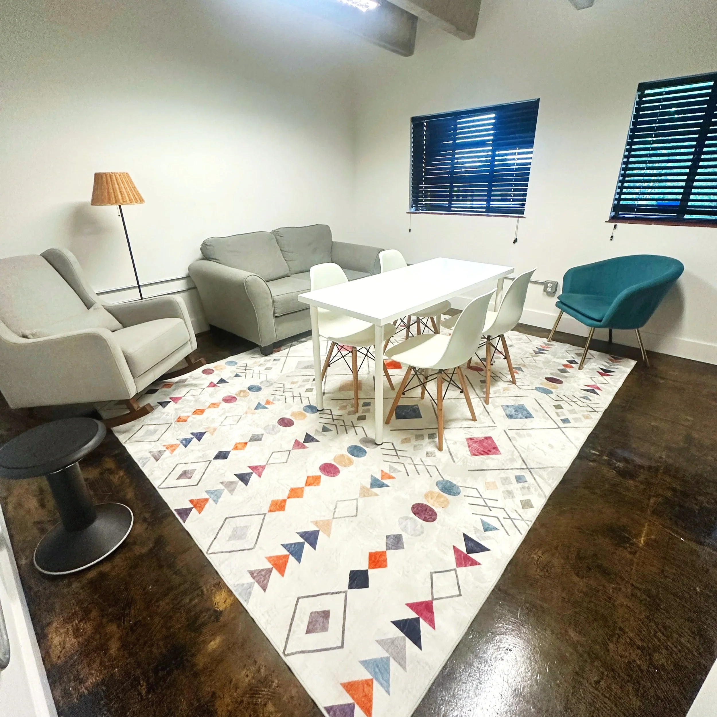 A living room with a colorful geometric patterned area rug, a light-colored sofa, a white coffee table, a white armchair, two teal armchairs, a wooden floor lamp, and two windows with dark blue blinds.