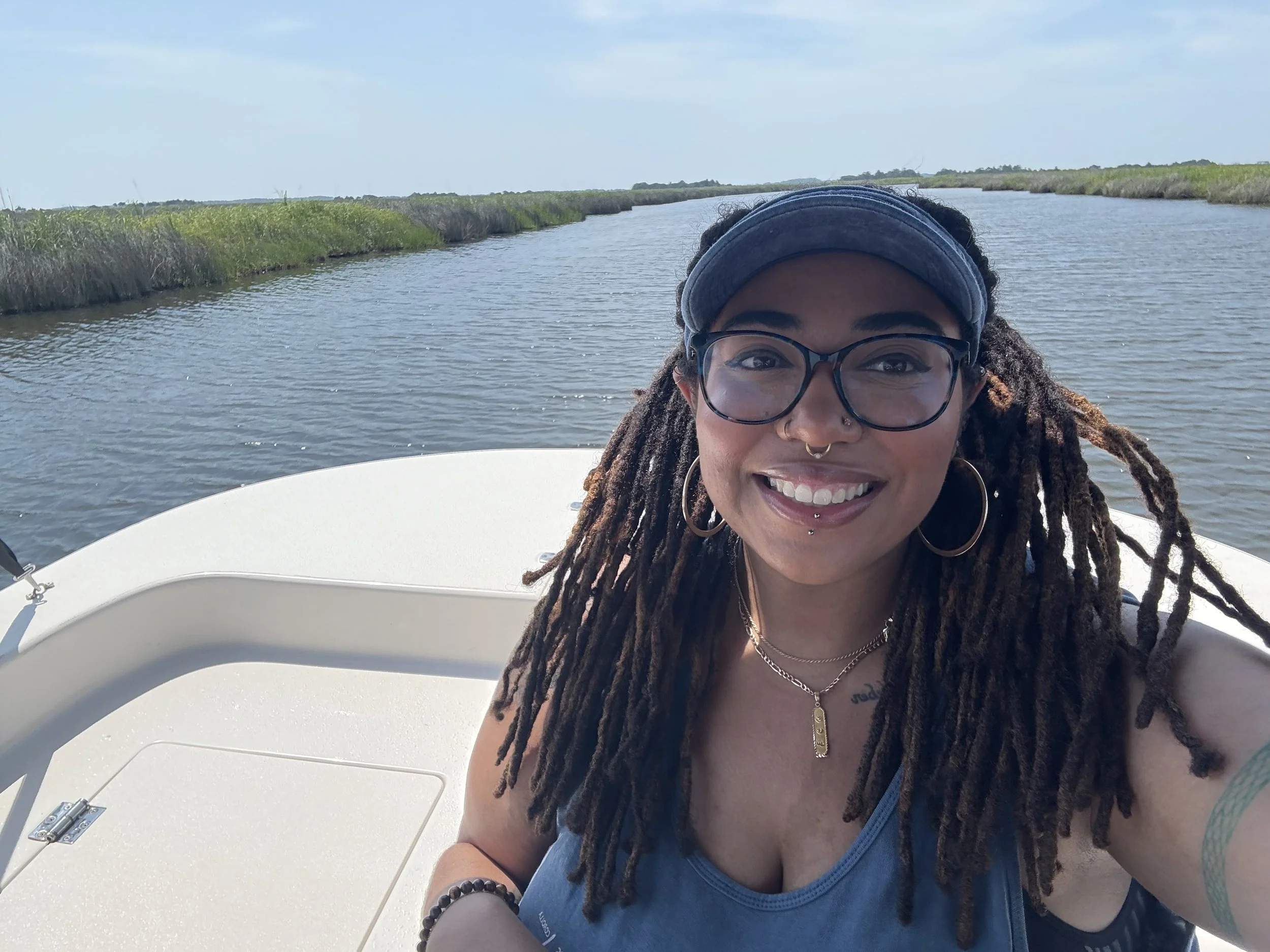 Young woman with dreadlocks wearing glasses, a headband, and jewelry, smiling on a boat with a waterway and marshland in the background.