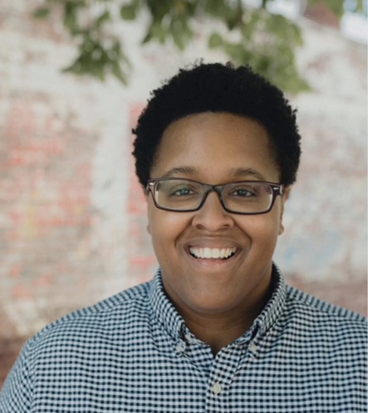 A smiling person with glasses and short curly hair, wearing a checkered shirt, standing outdoors with a blurred brick wall and some green leaves in the background.