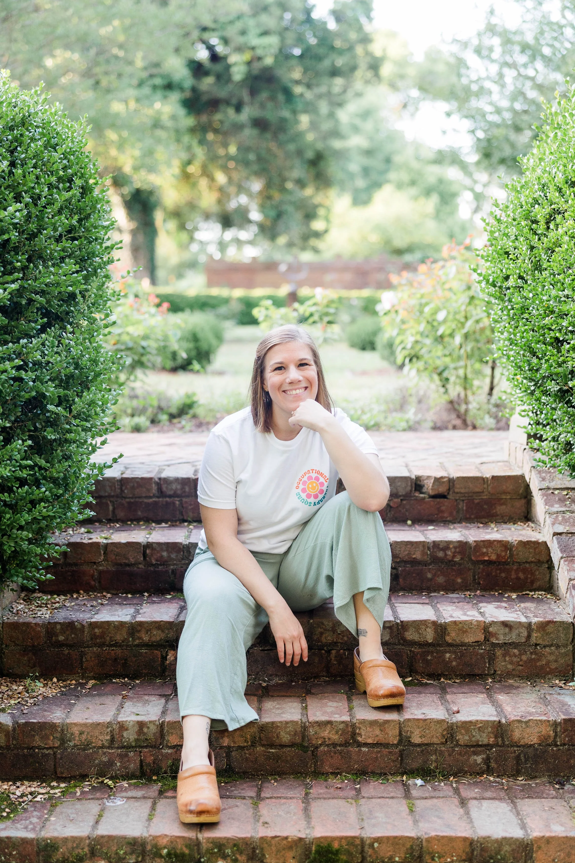 A smiling young woman with shoulder-length brown hair sitting on brick steps in a garden, surrounded by green hedges and trees, wearing a white t-shirt, light green pants, and brown shoes.