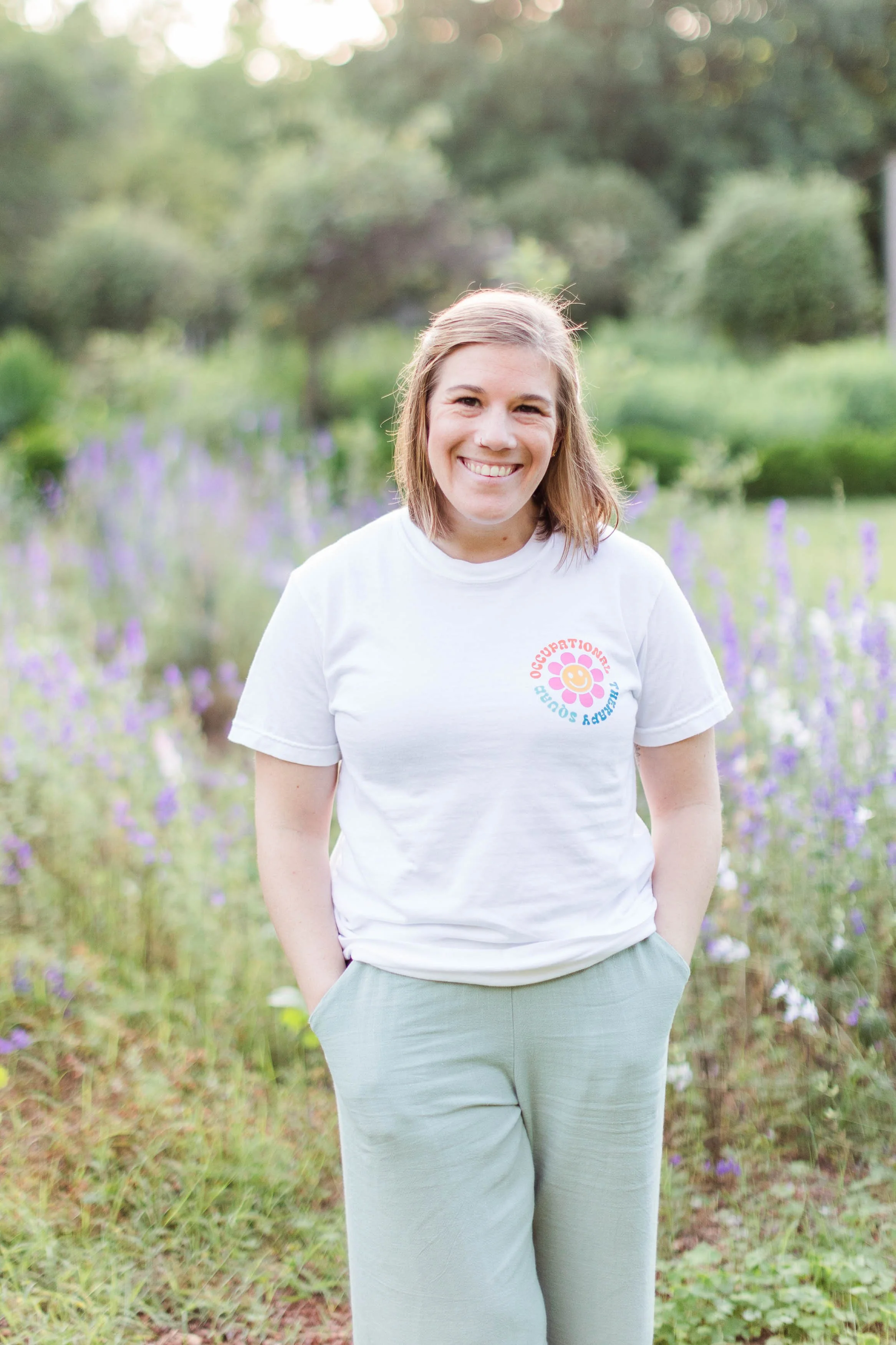 A woman with shoulder-length light brown hair, smiling, wearing a white t-shirt with a colorful logo on it, standing outdoors in front of a blurred background of green trees and purple flowers.