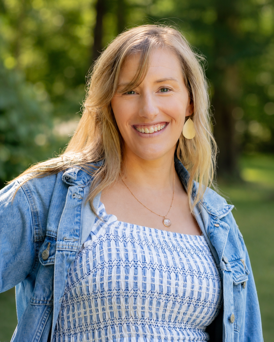 A woman smiling outdoors, wearing a denim jacket, a patterned blouse, and jewelry, with green foliage in the background.