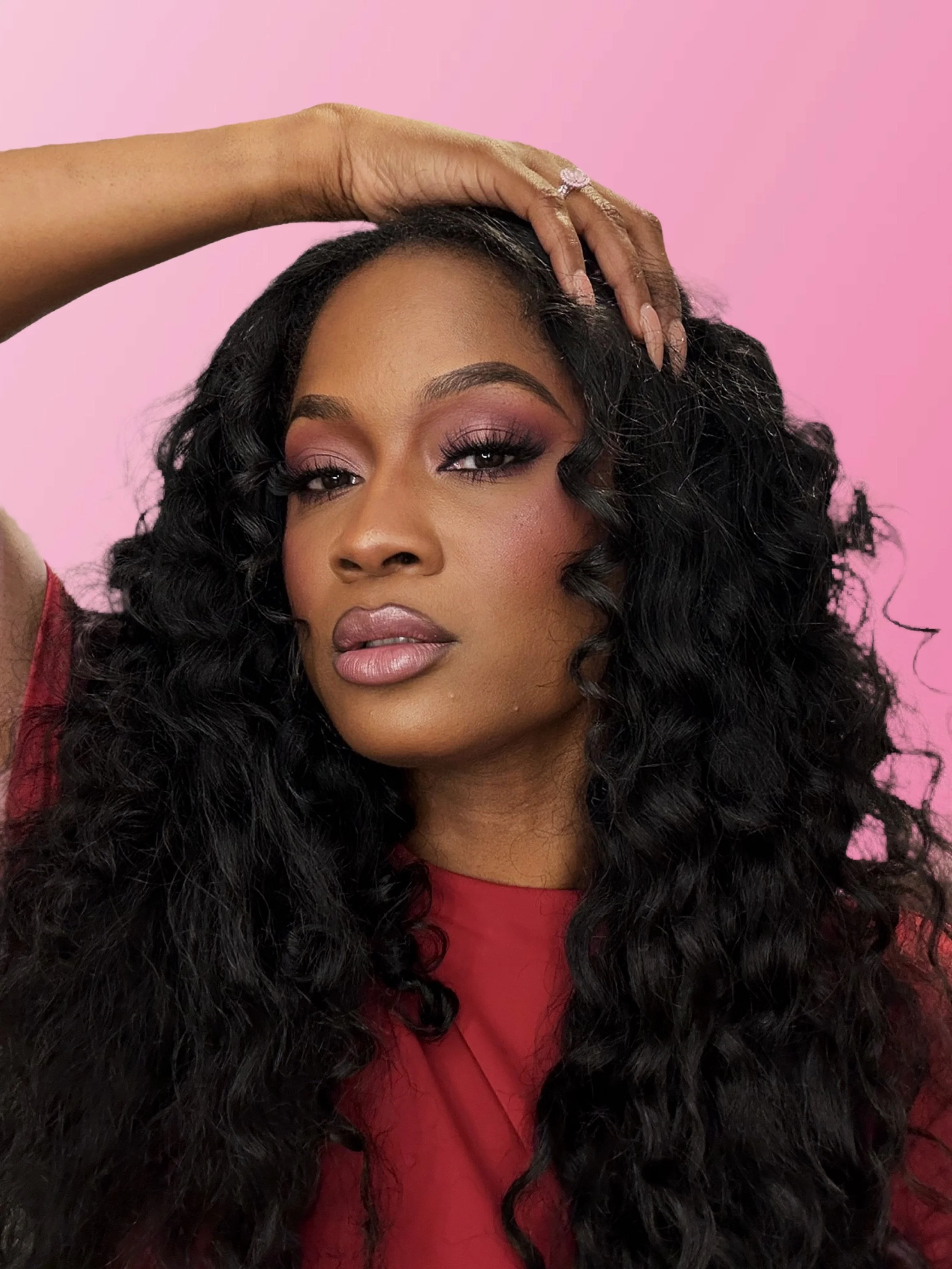 Close-up of a woman with long black curly hair, wearing makeup with pink and purple eyeshadow and pink lipstick, against a pink background, holding her hand on her head and showing a ring on her finger.