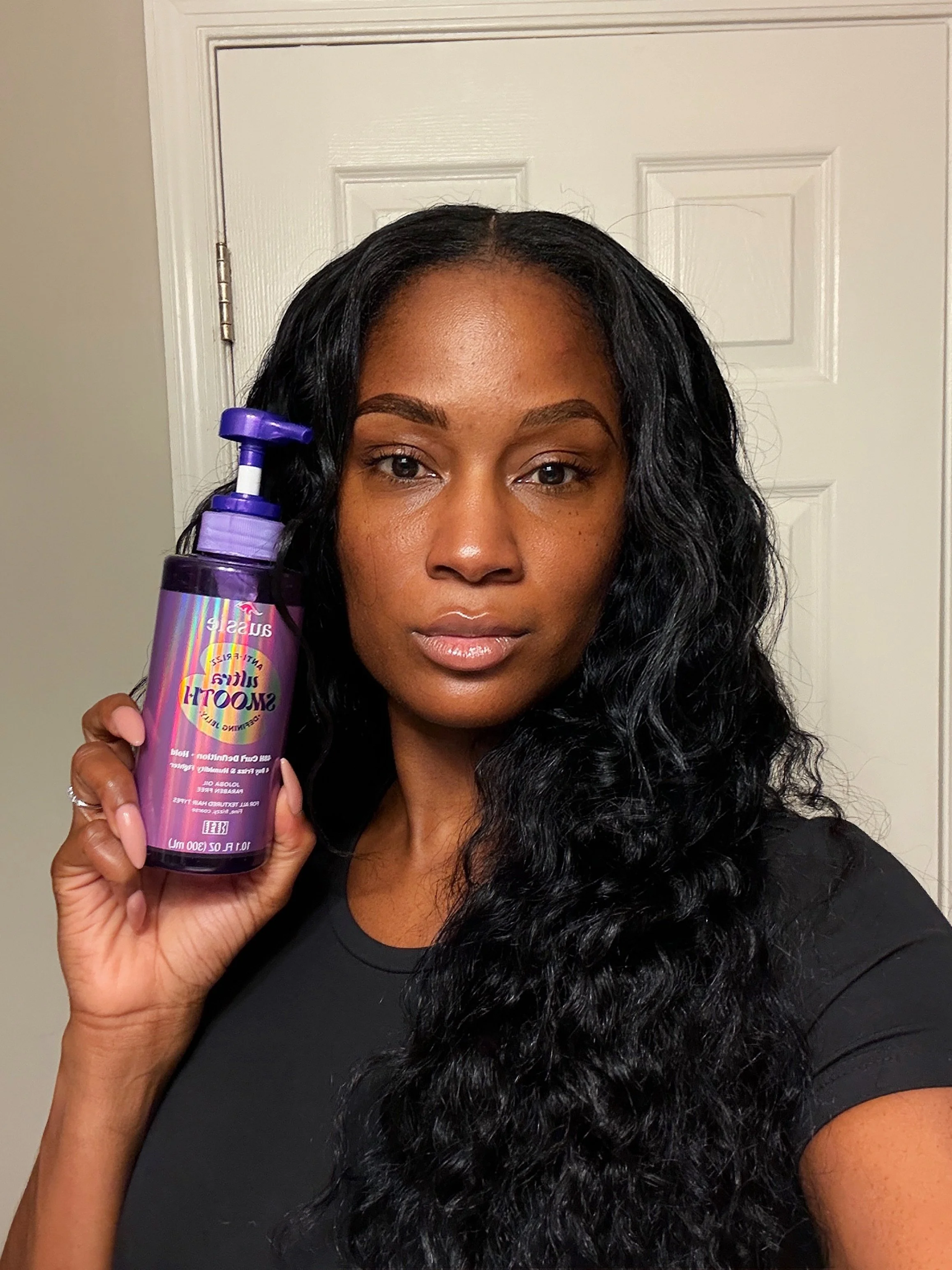 A woman with long, curly black hair holding a purple bottle of shampoo in front of a white door.