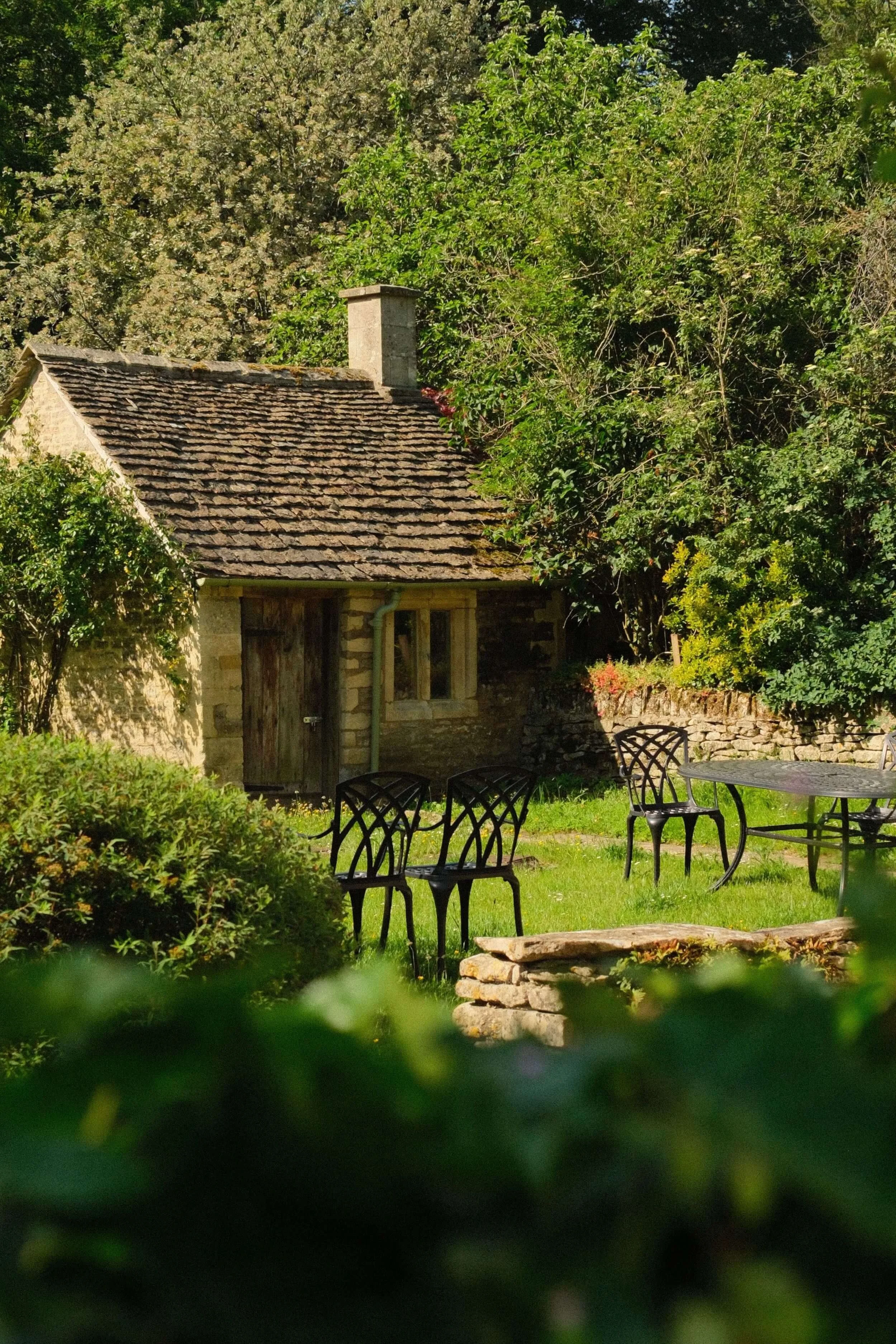 Stone cottage with a slate roof and chimney surrounded by lush garden greenery and outdoor café furniture