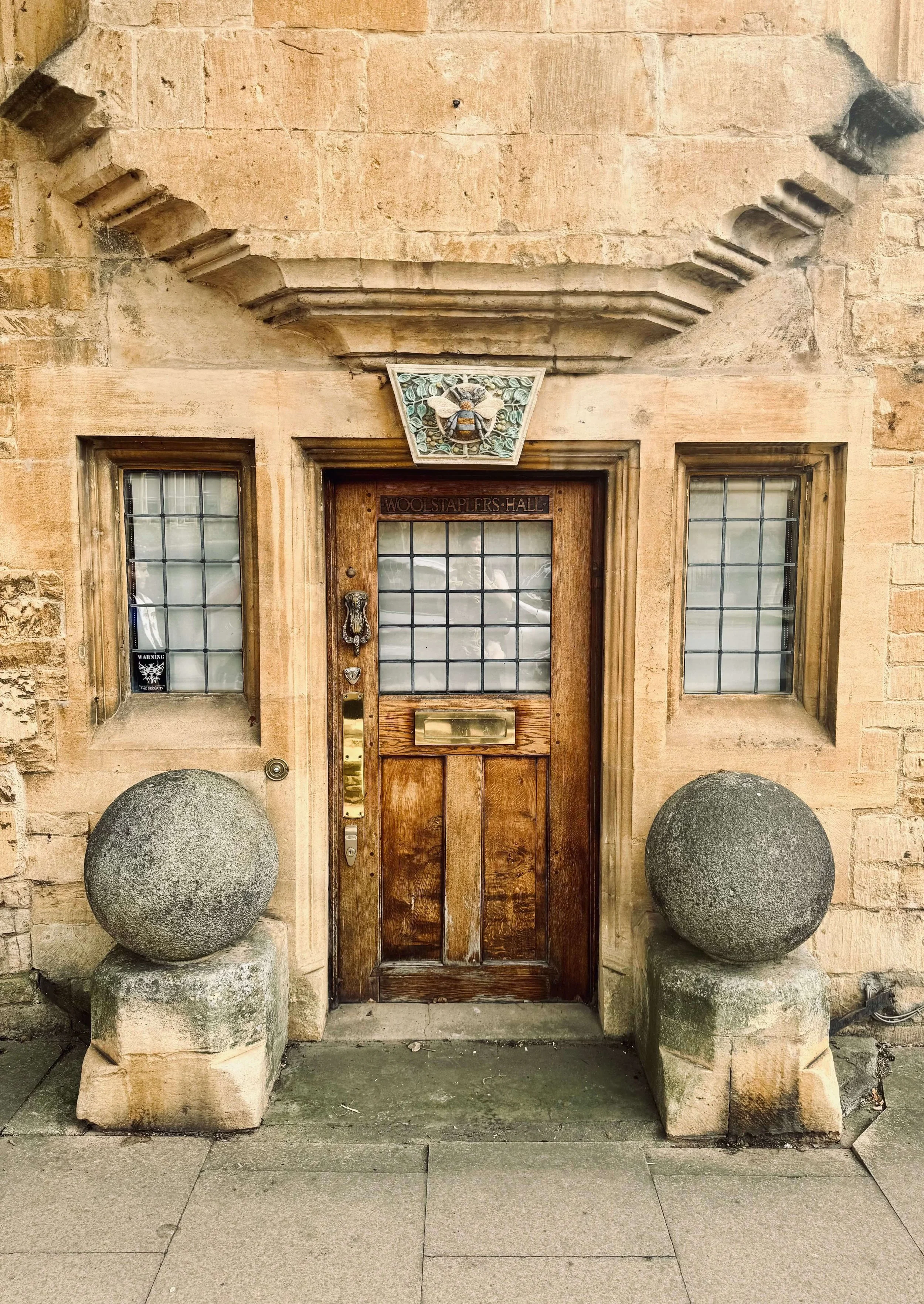 Historic stone Woolstaplers' Hall entrance with a carved crest, wooden door, leaded-glass windows, and stone spheres.