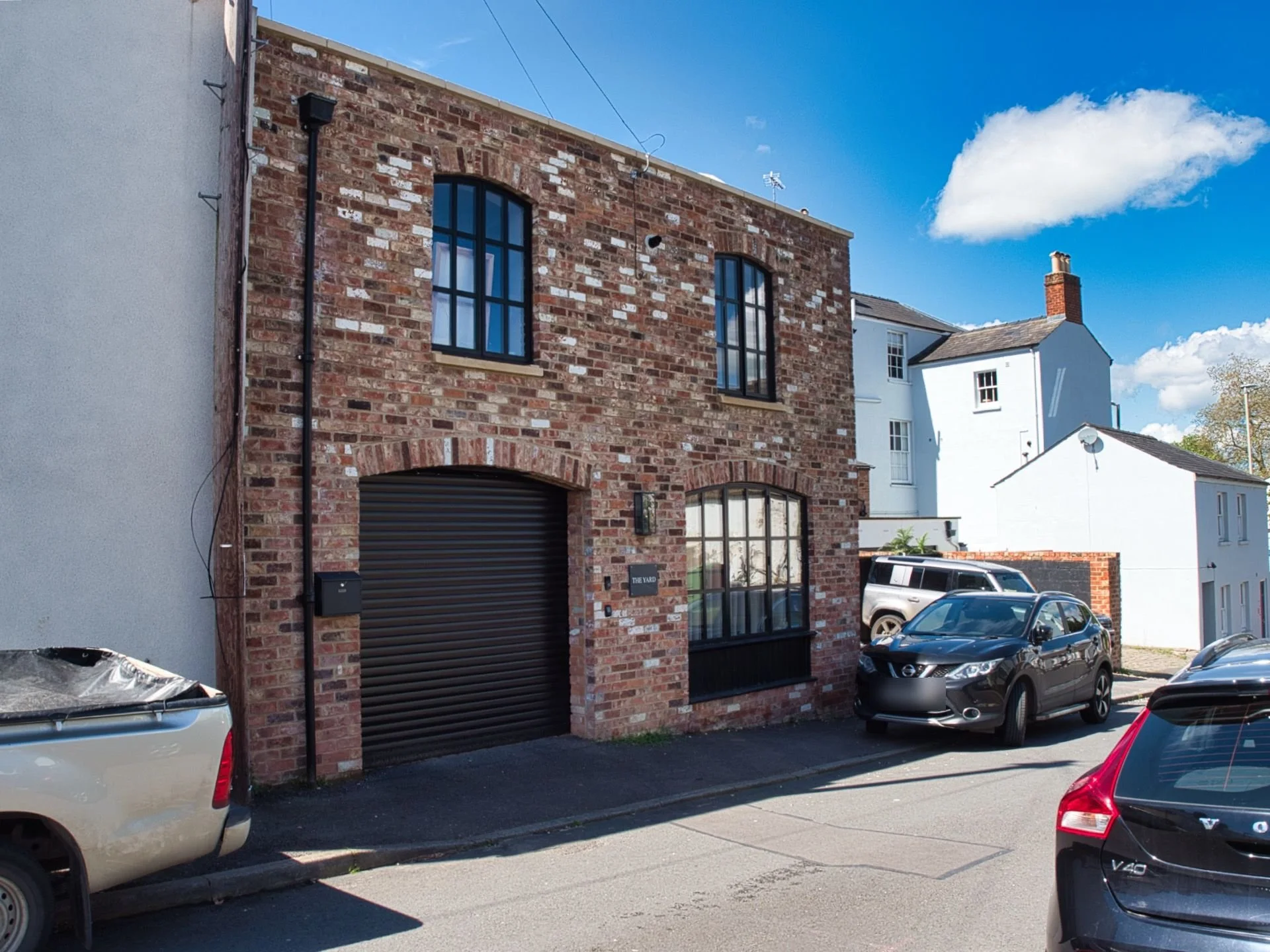 Brick building with arched windows and garage door on a sunny residential street, surrounded by parked cars.