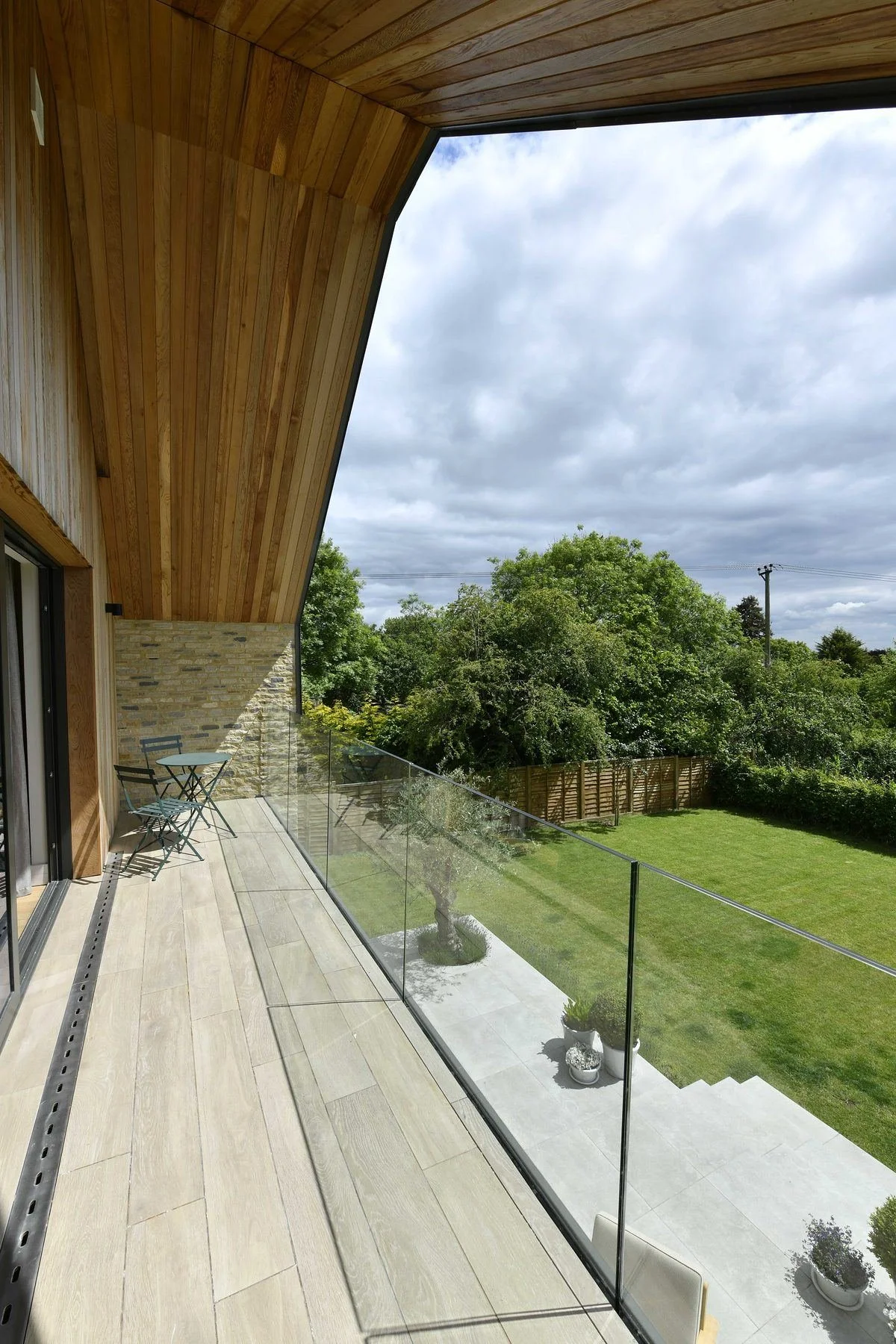 Modern home balcony with glass railing, wooden overhang, outdoor table and chairs, landscaped garden, and cloudy sky