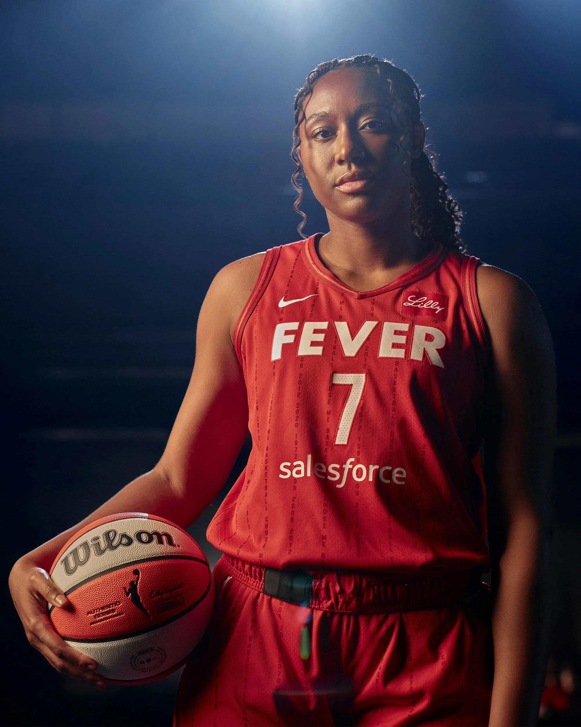 Indiana Fever basketball player wearing a red No. 7 jersey poses with a Wilson basketball under dramatic lighting.