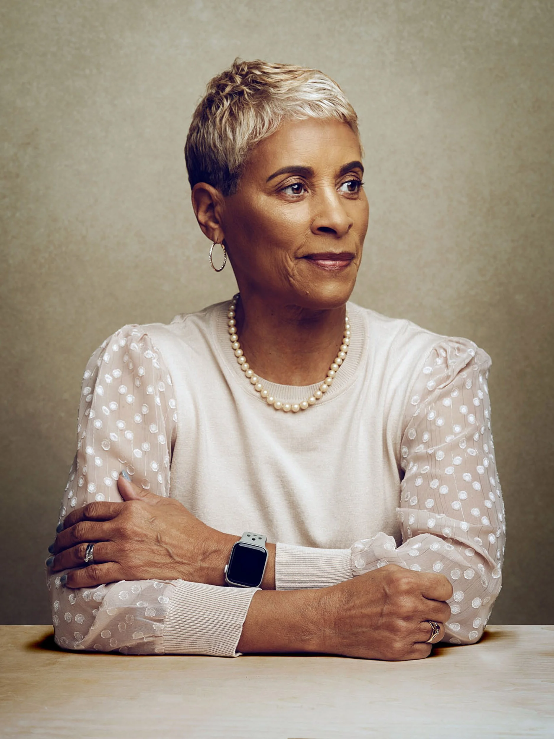 Portrait of an older woman with short gray hair wearing a pearl necklace and white textured blouse, seated with folded arms.