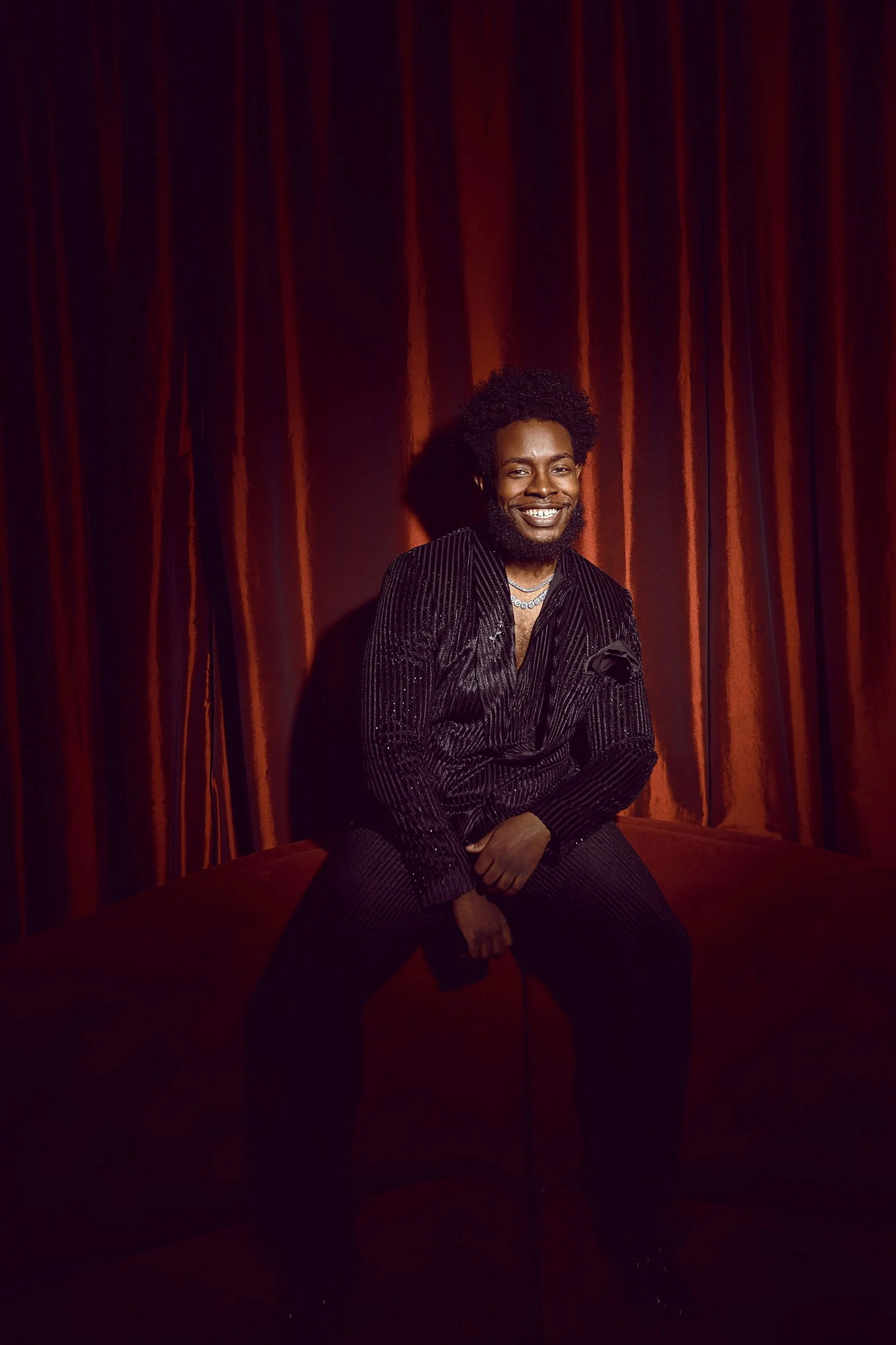 Smiling man in a black pinstripe suit seated in front of red velvet curtains