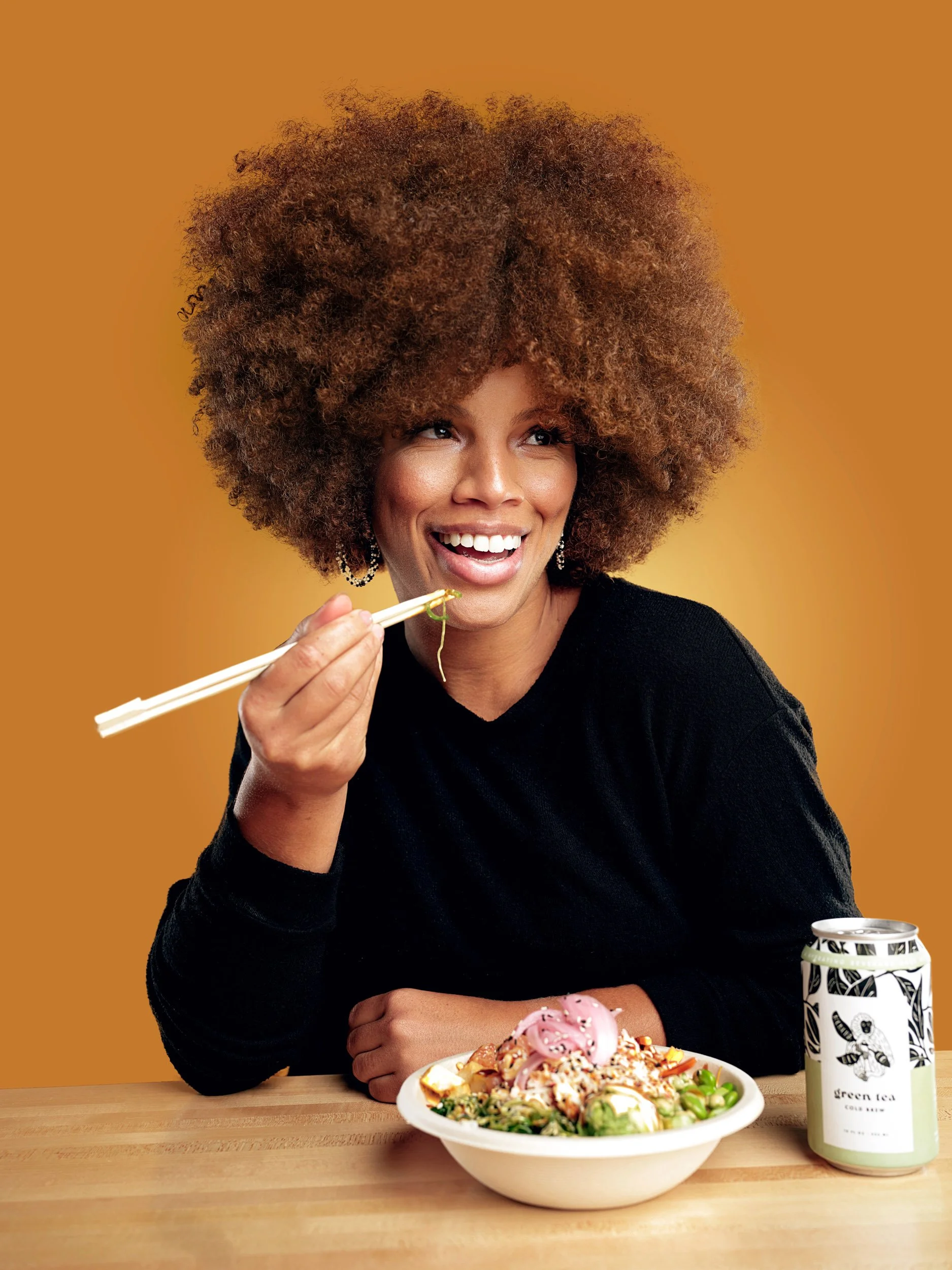 Smiling woman eating noodle salad with chopsticks beside a bowl of food and green tea