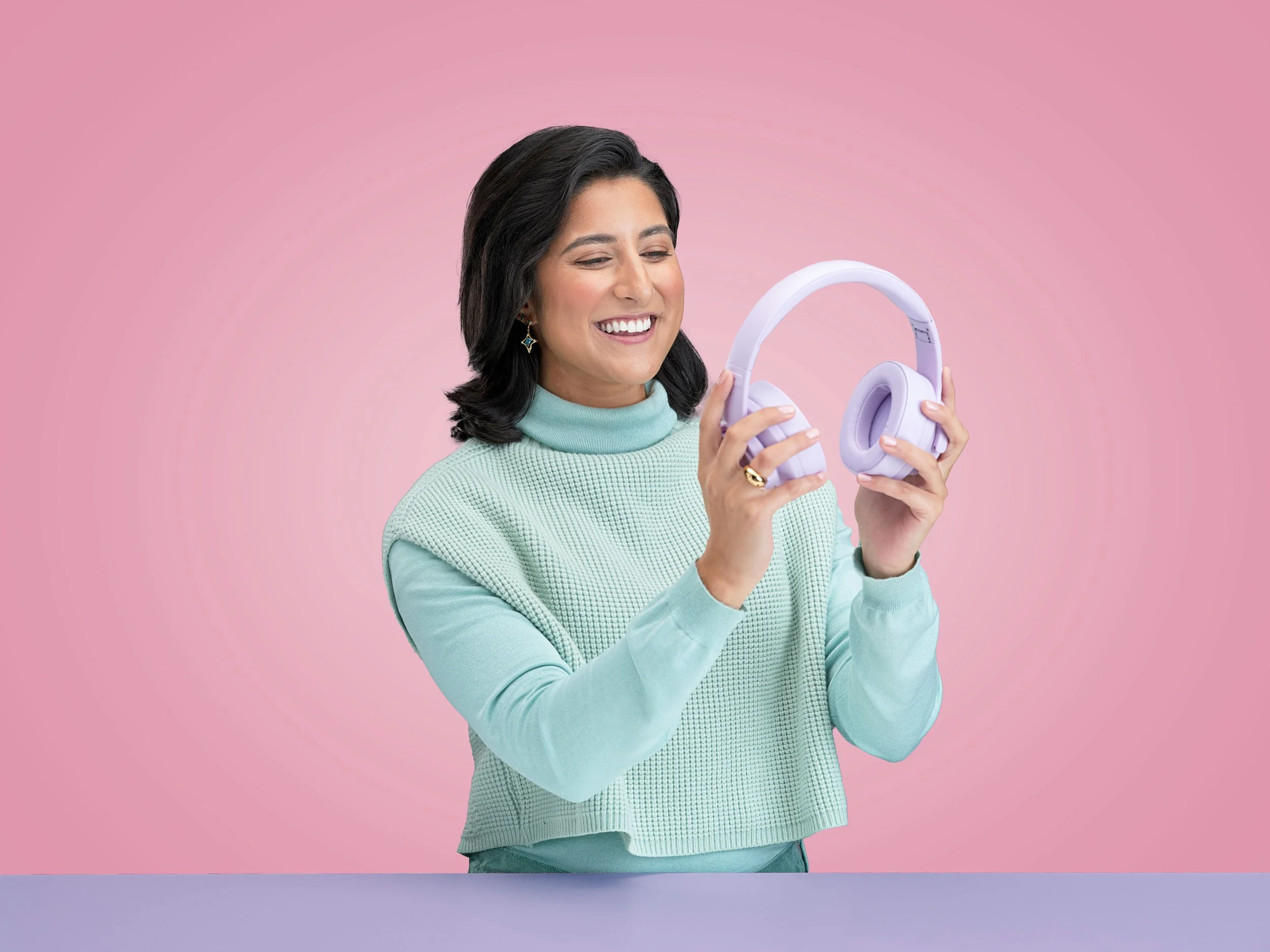 Smiling woman holding lavender over-ear headphones against a pink background