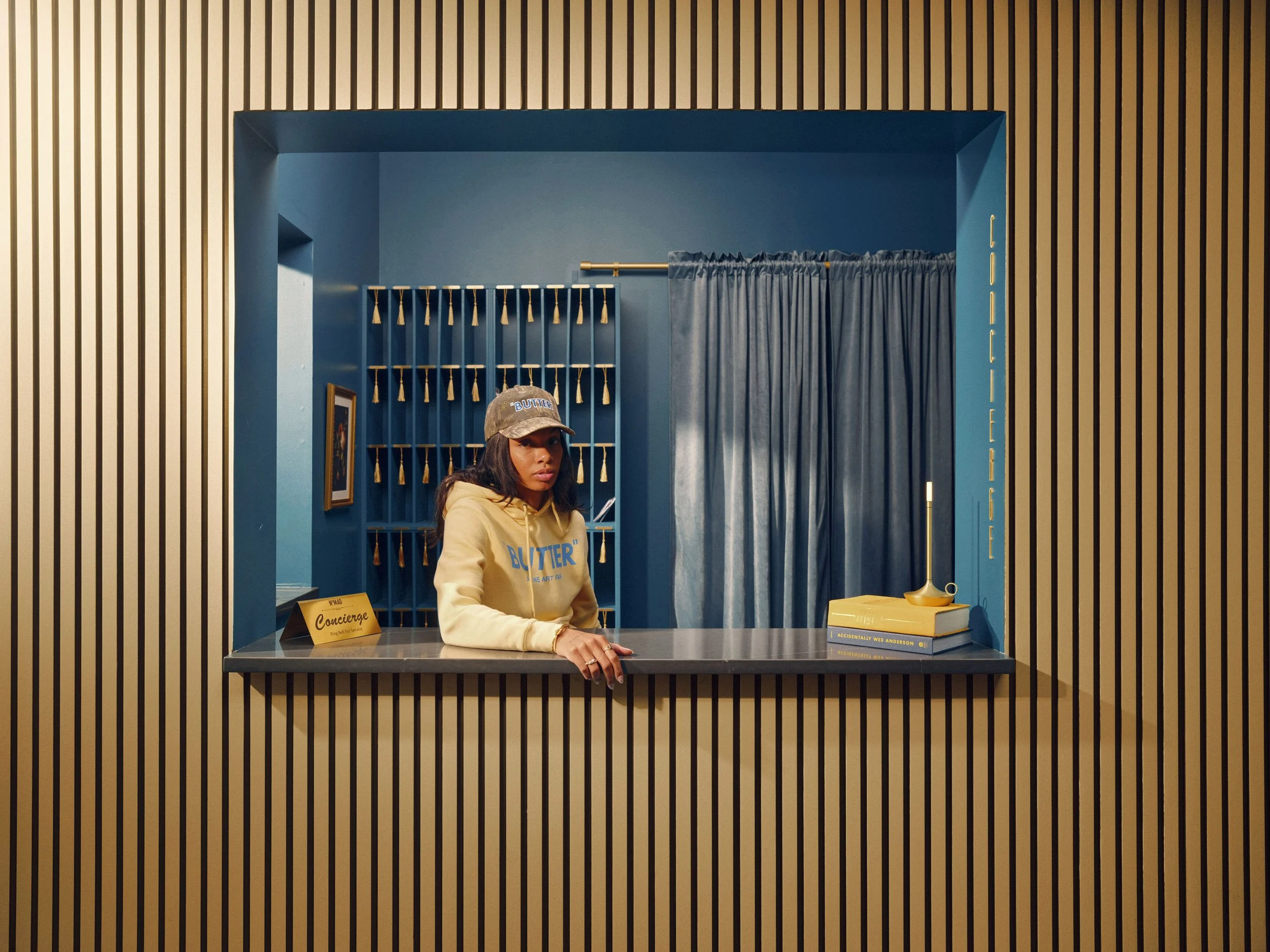 Woman in a BUTTER hoodie and cap behind a concierge desk in a blue-and-gold hotel lobby