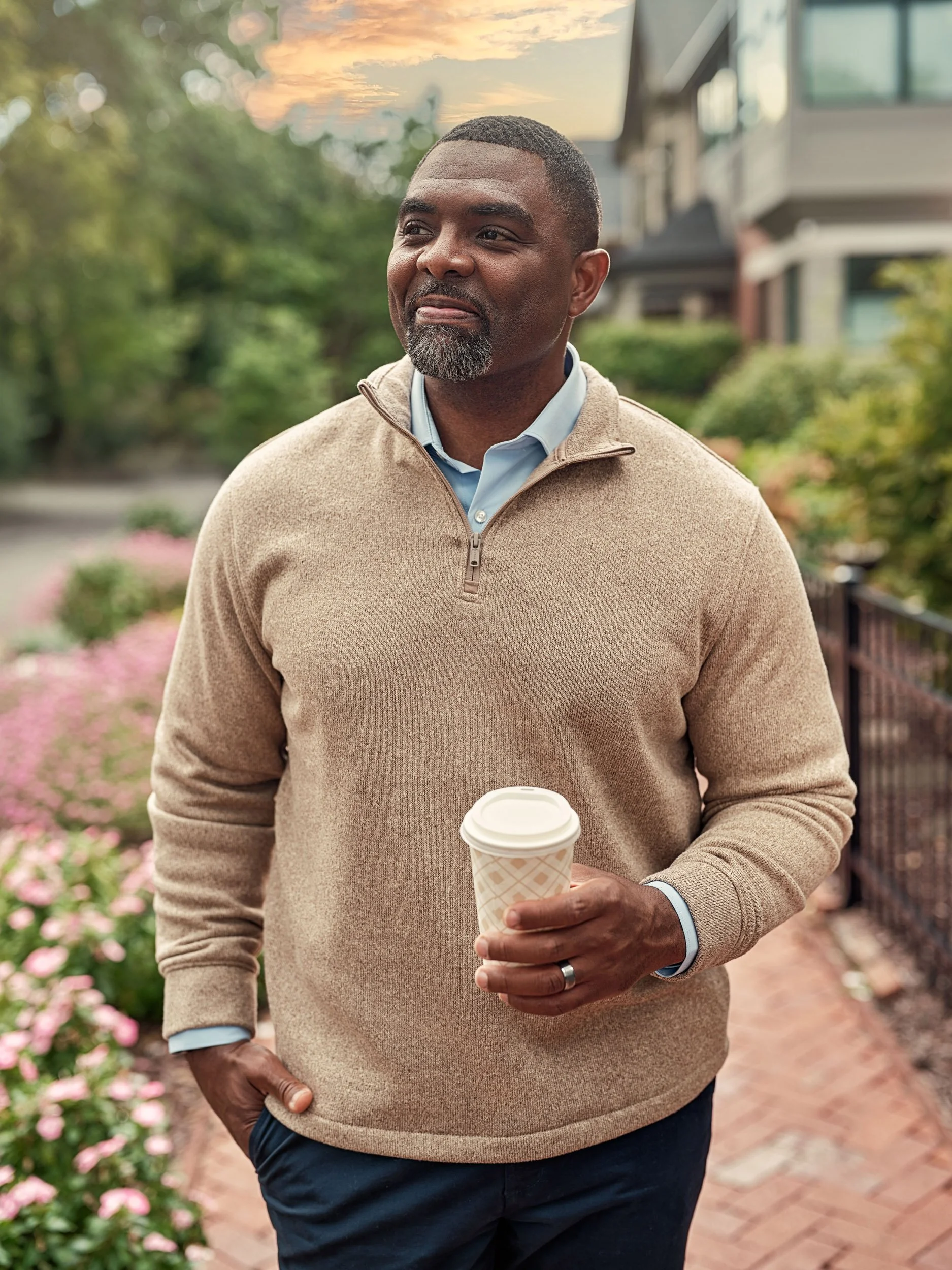 Smiling Black man in a beige sweater holding a takeaway coffee cup outdoors in a garden courtyard