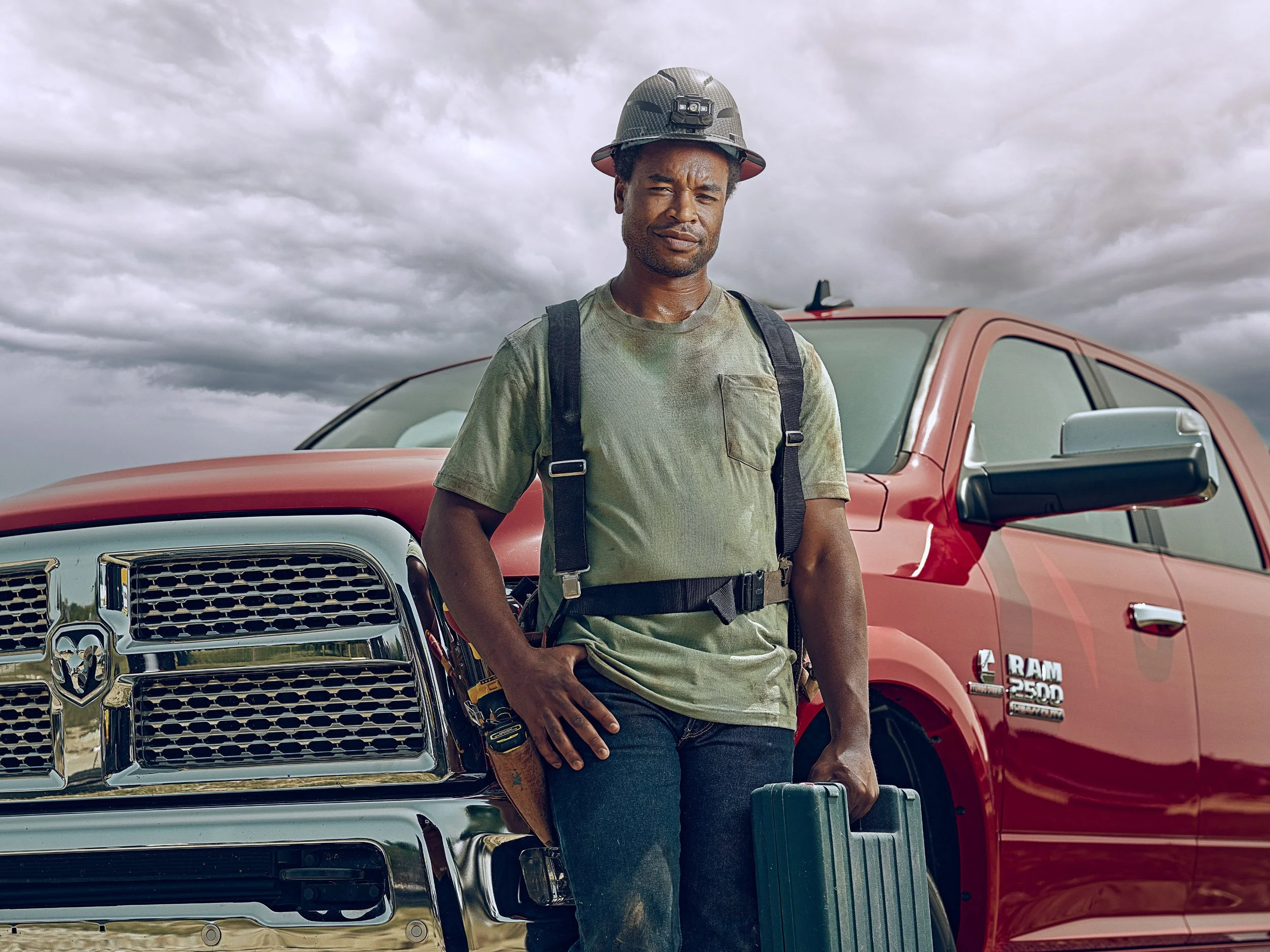 Black male mechanic wearing a hard hat and tool belt standing between two pickup trucks under a cloudy sky
