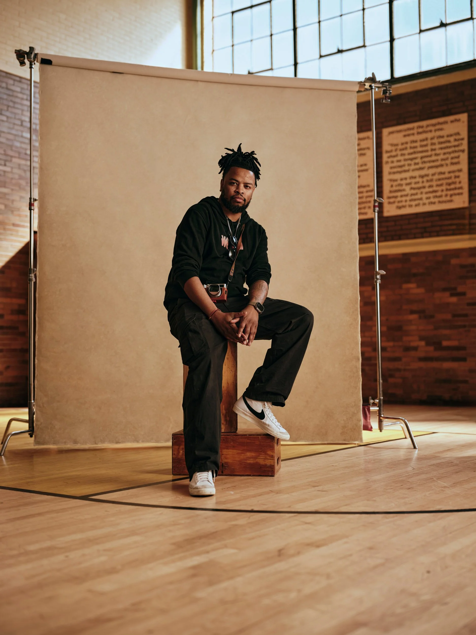 Man in black clothing sitting on a wooden box during a studio photoshoot with a camera and backdrop