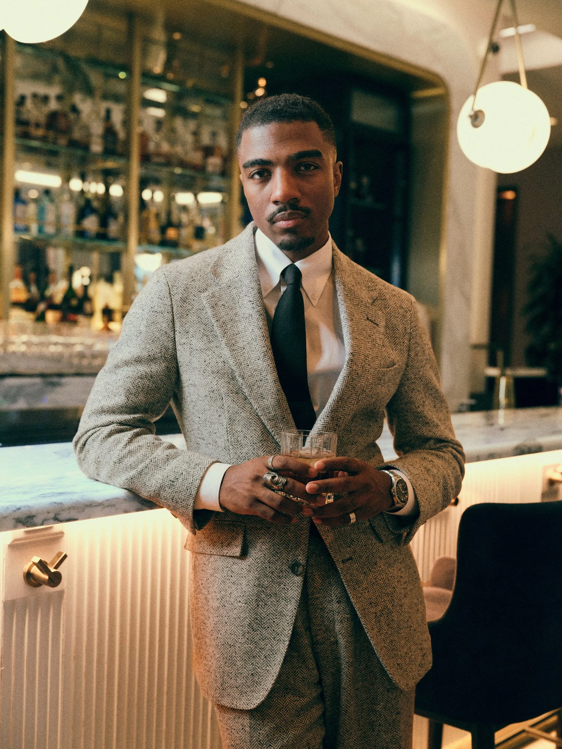 Well-dressed man in a herringbone suit and tie holding a drink at an upscale bar