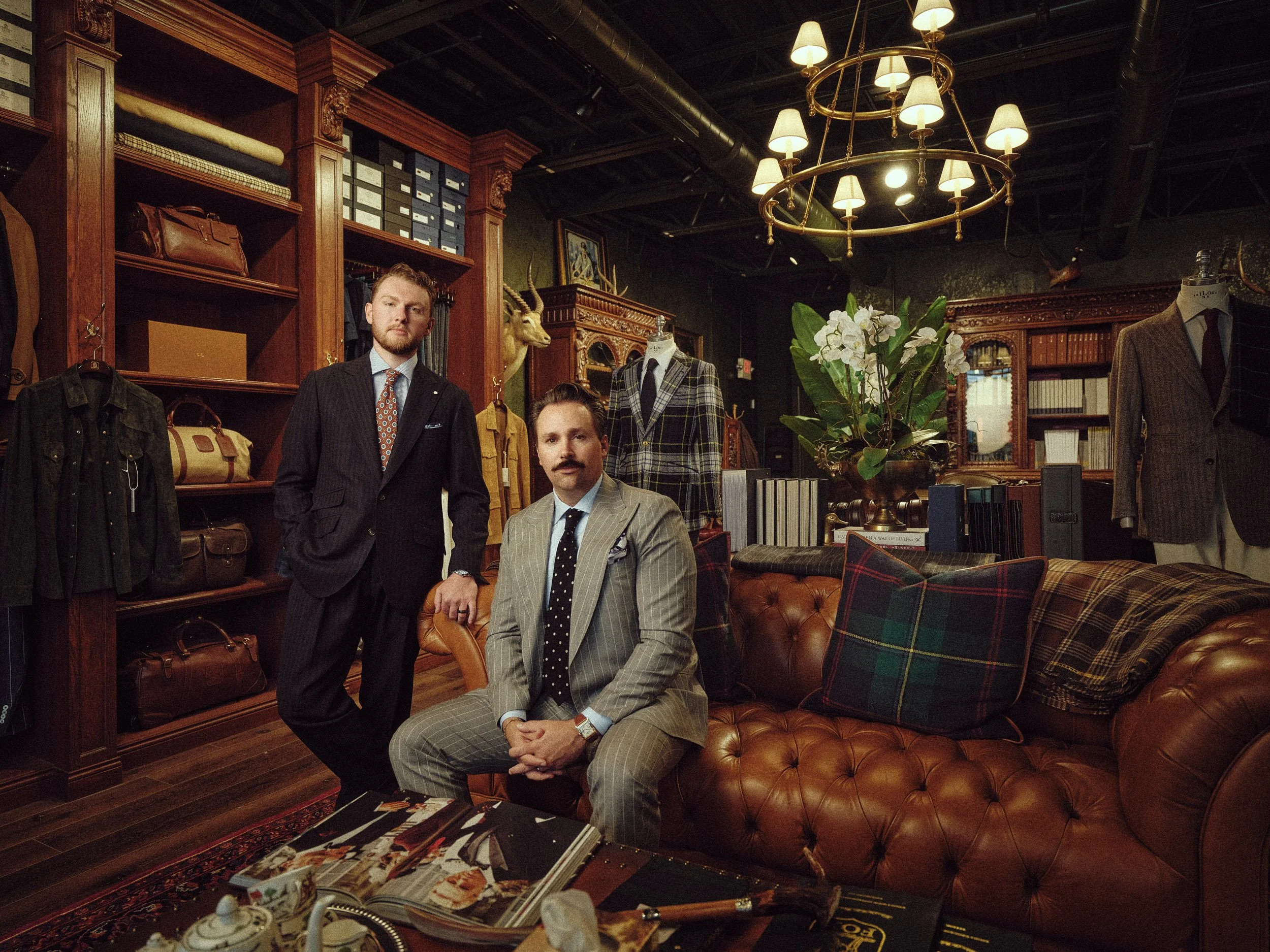 Three men in tailored suits and mannequins in a vintage luxury menswear boutique with leather bags, books, a chandelier, and a tufted leather sofa Carmel Indiana
