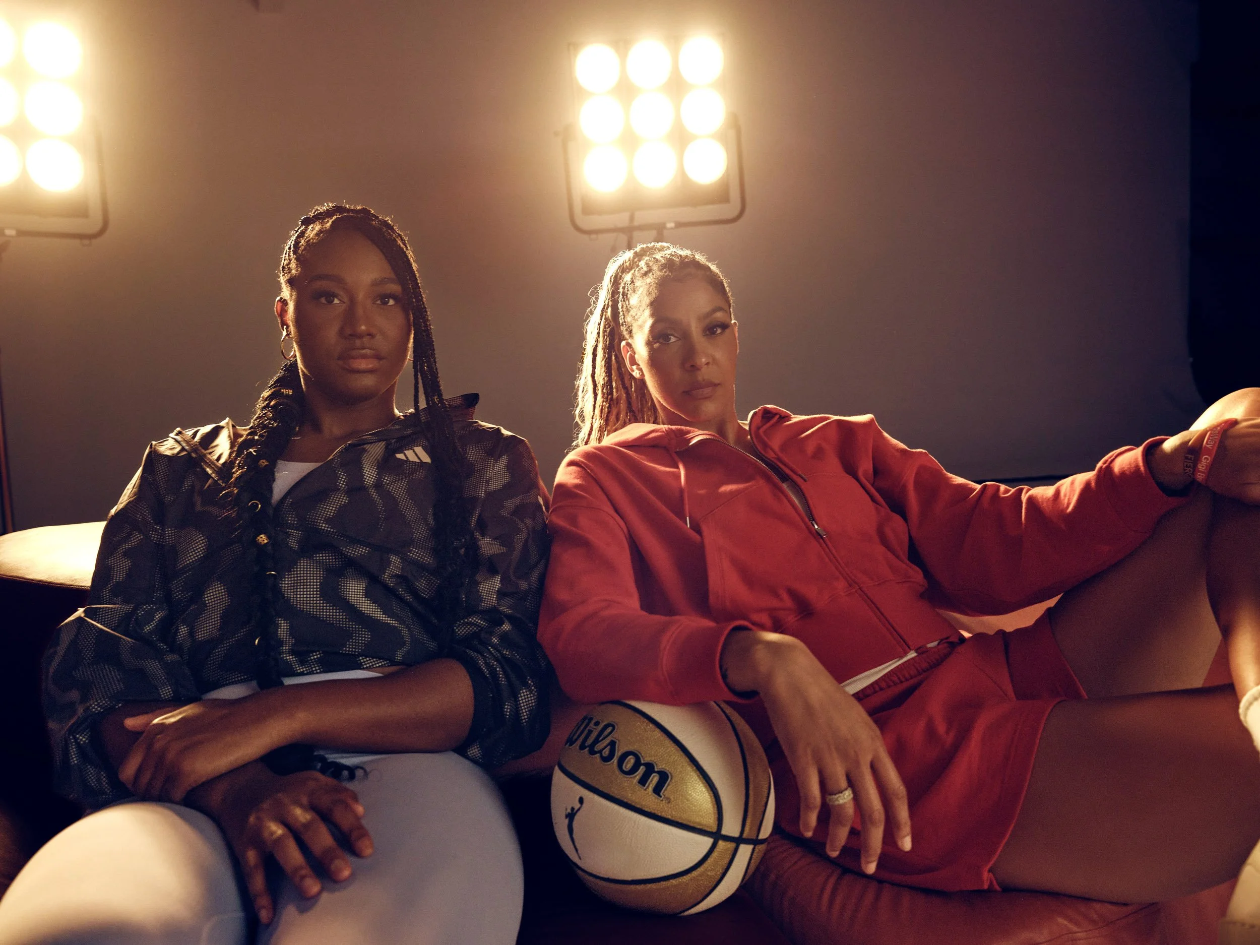 Two women in athletic clothing sit courtside with a basketball under bright arena lights.