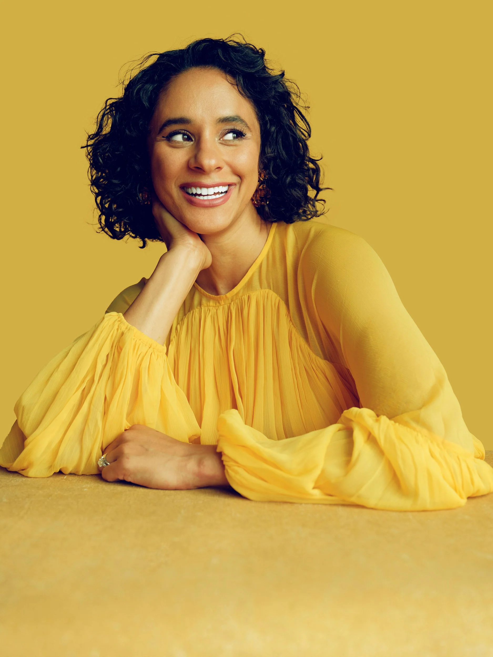 Smiling woman with curly dark hair wearing a flowing yellow dress against a mustard-yellow background