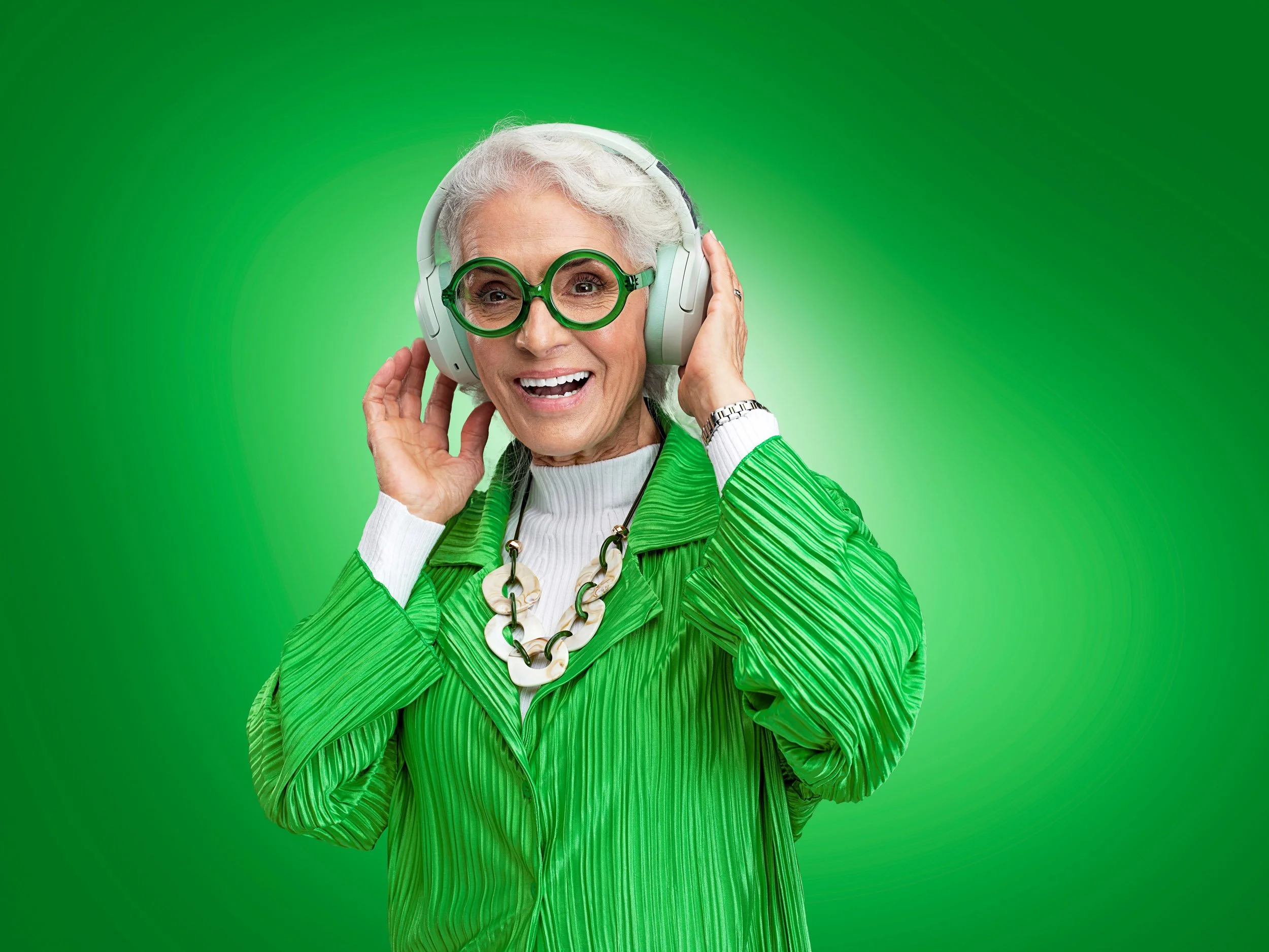 Smiling older woman wearing green glasses, a green pleated outfit, and white over-ear headphones against a bright green background