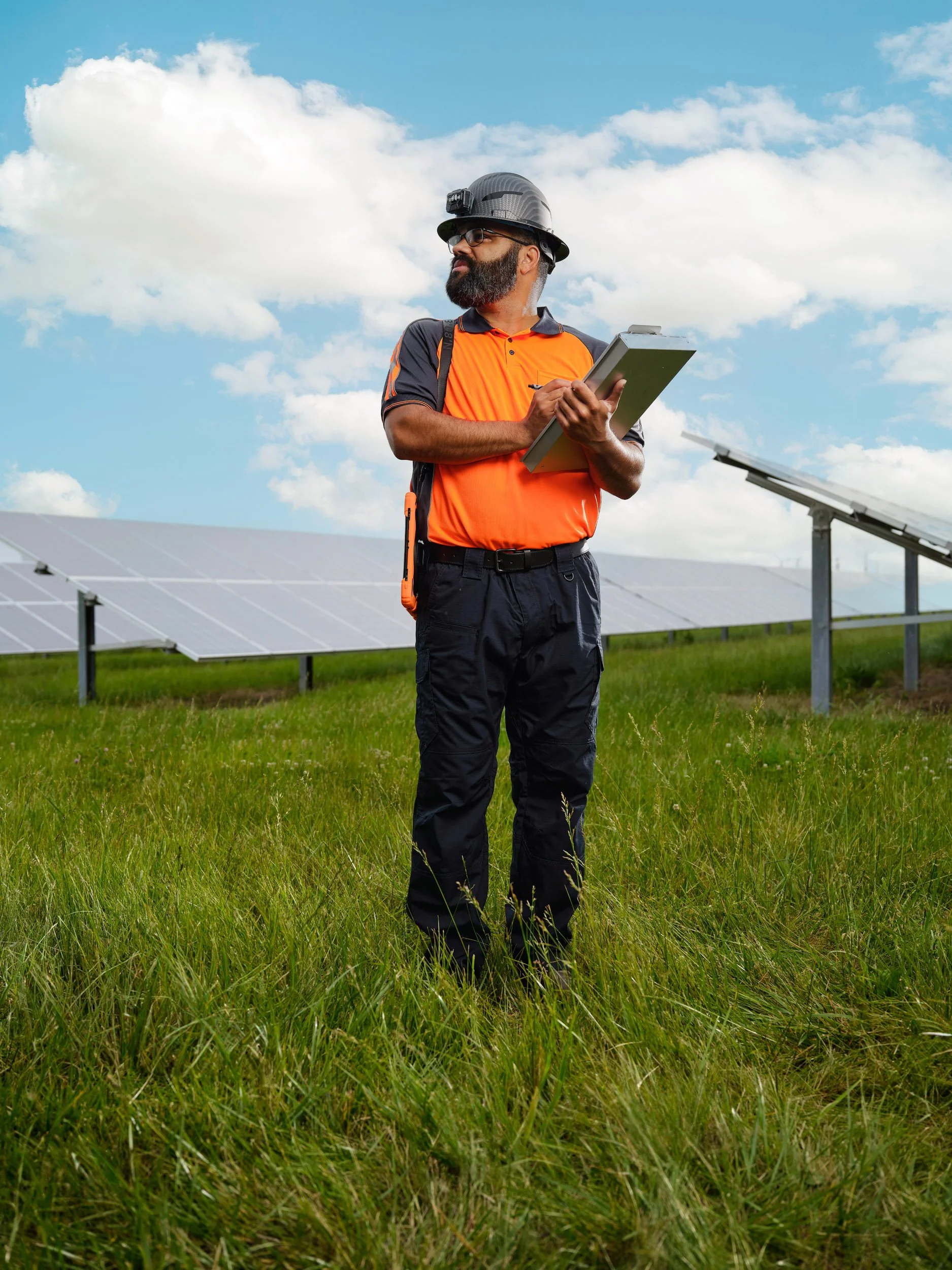 Solar technician in a safety helmet inspecting solar panels with a clipboard in a grassy field