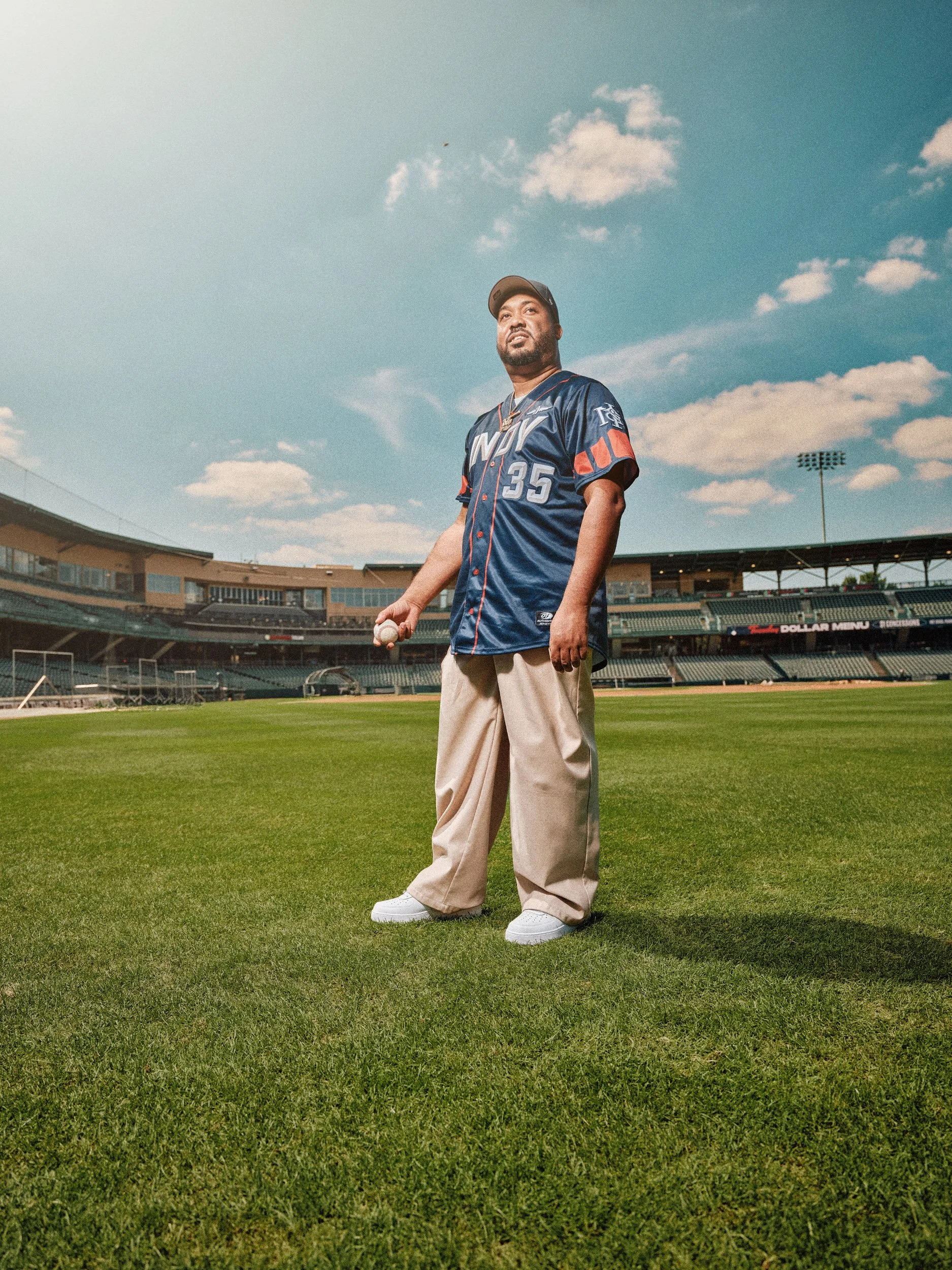 B Swift Radio Personality wearing a navy jersey with number 35 holds a baseball on a stadium field under a blue sky for pattern Magazine