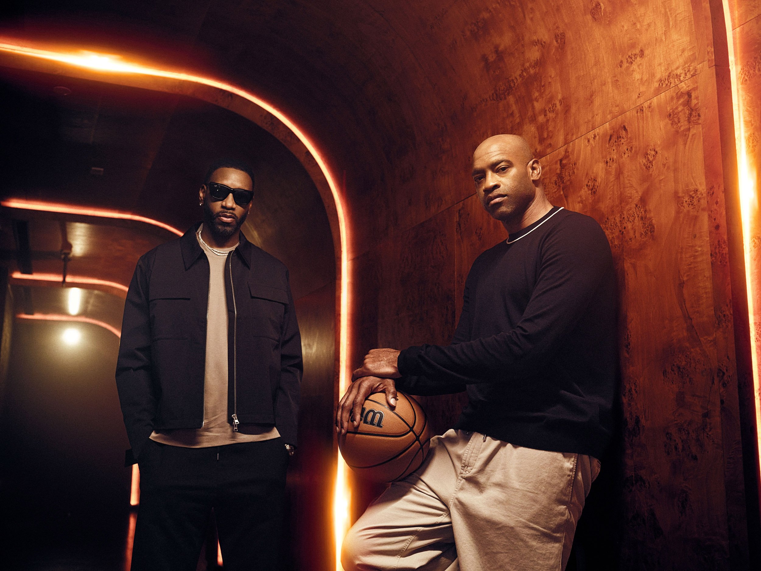 Vince Carter and Tracy Mcgrady Two men posing in a warmly lit wood-paneled room, with one holding a basketball and the other wearing sunglasses.