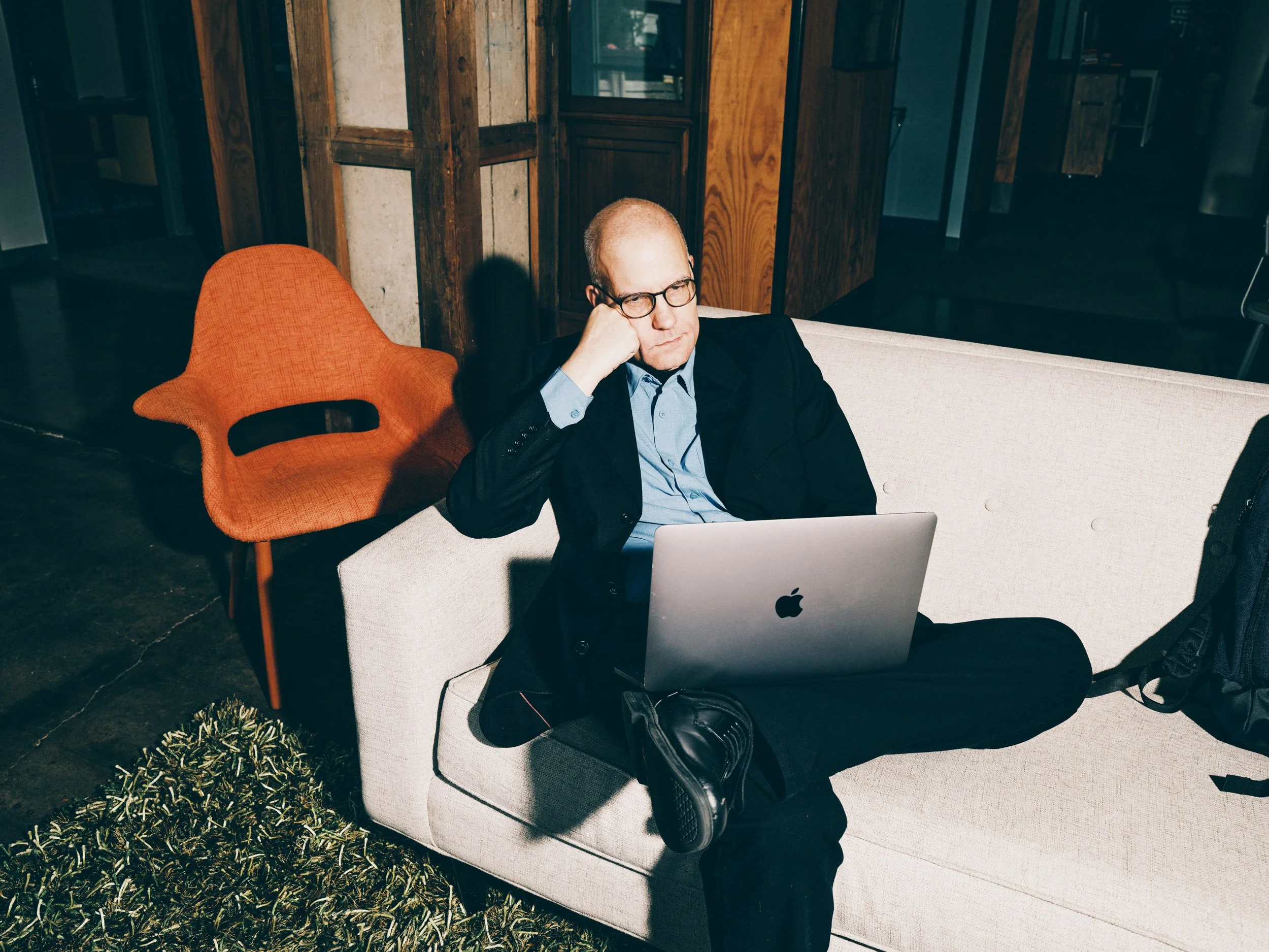 Bald man wearing glasses works on a laptop while sitting on a white couch in a rustic interior.