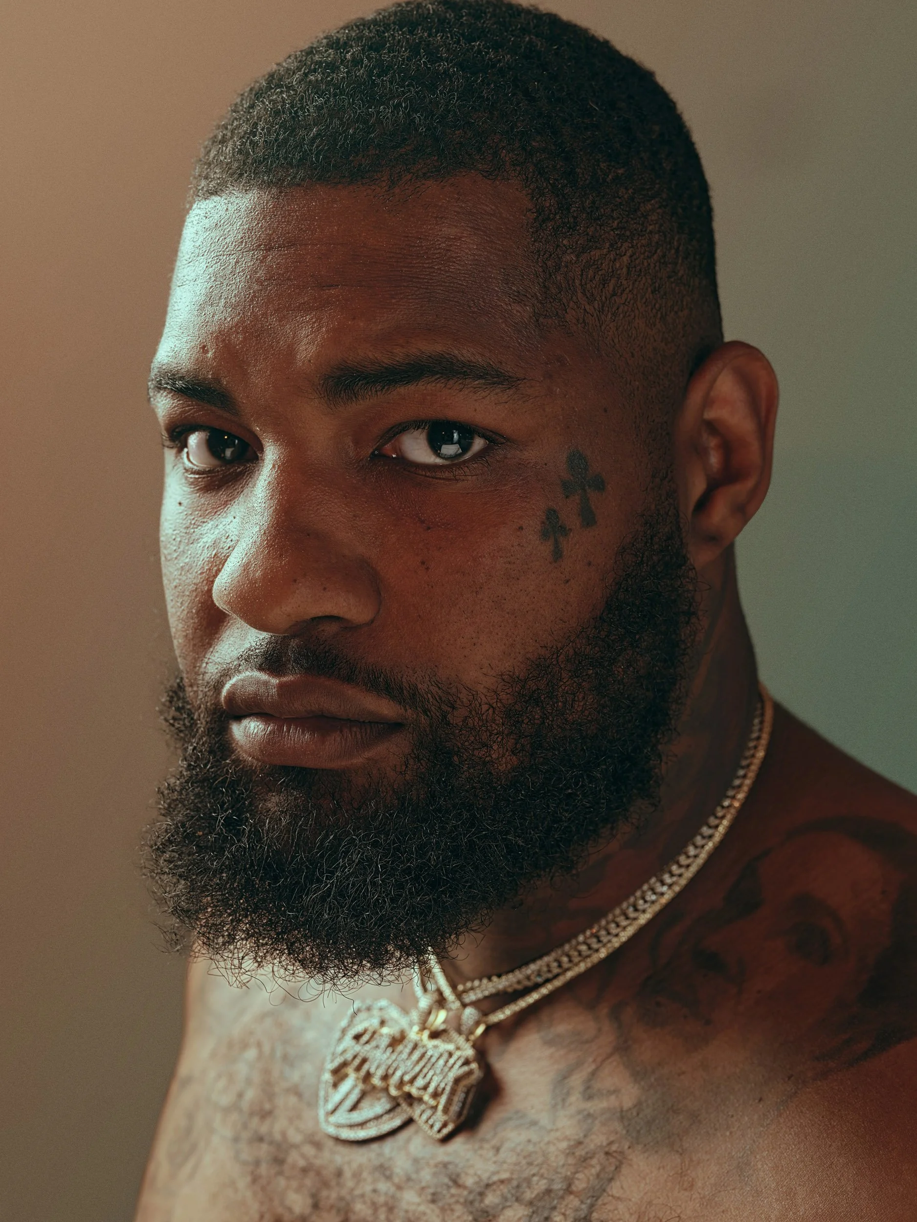 Close-up portrait of a bearded Black man with a short haircut, facial tattoos, neck tattoos, and layered gold chains.