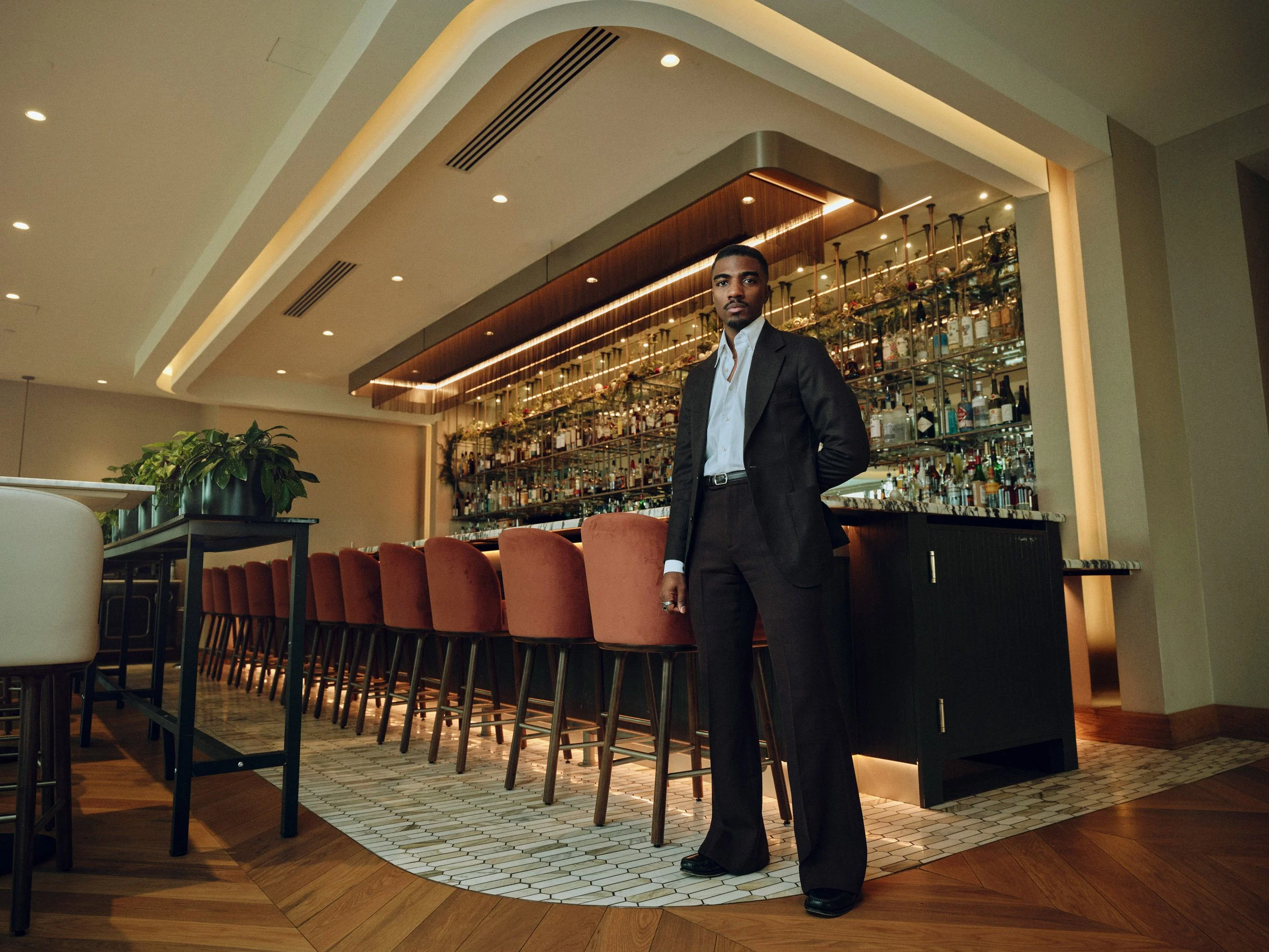 Man in a black suit standing inside an upscale bar with a wall of liquor bottles and bar stools