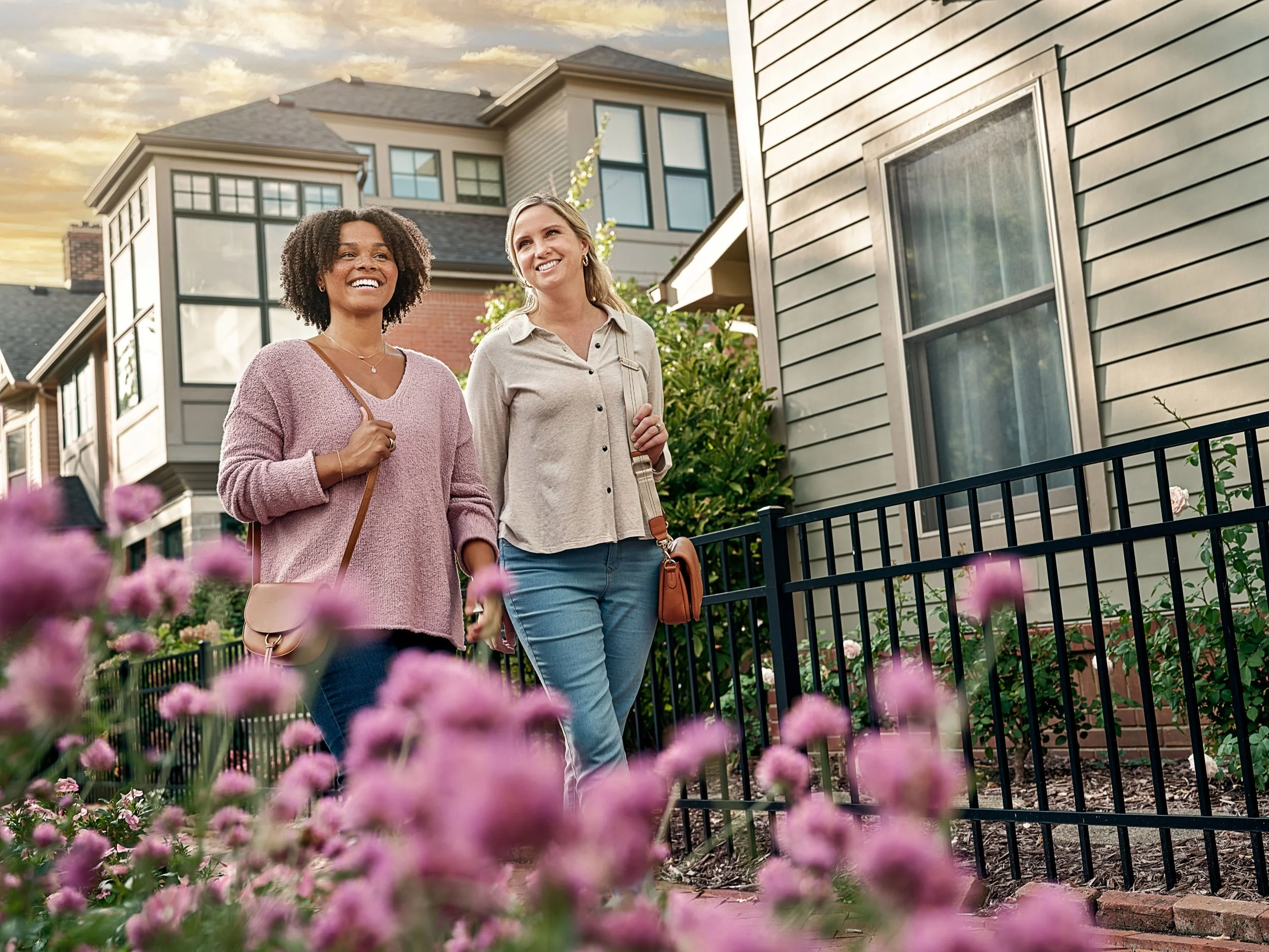 Two smiling women walking through a flower-filled residential neighborhood beside a modern home.