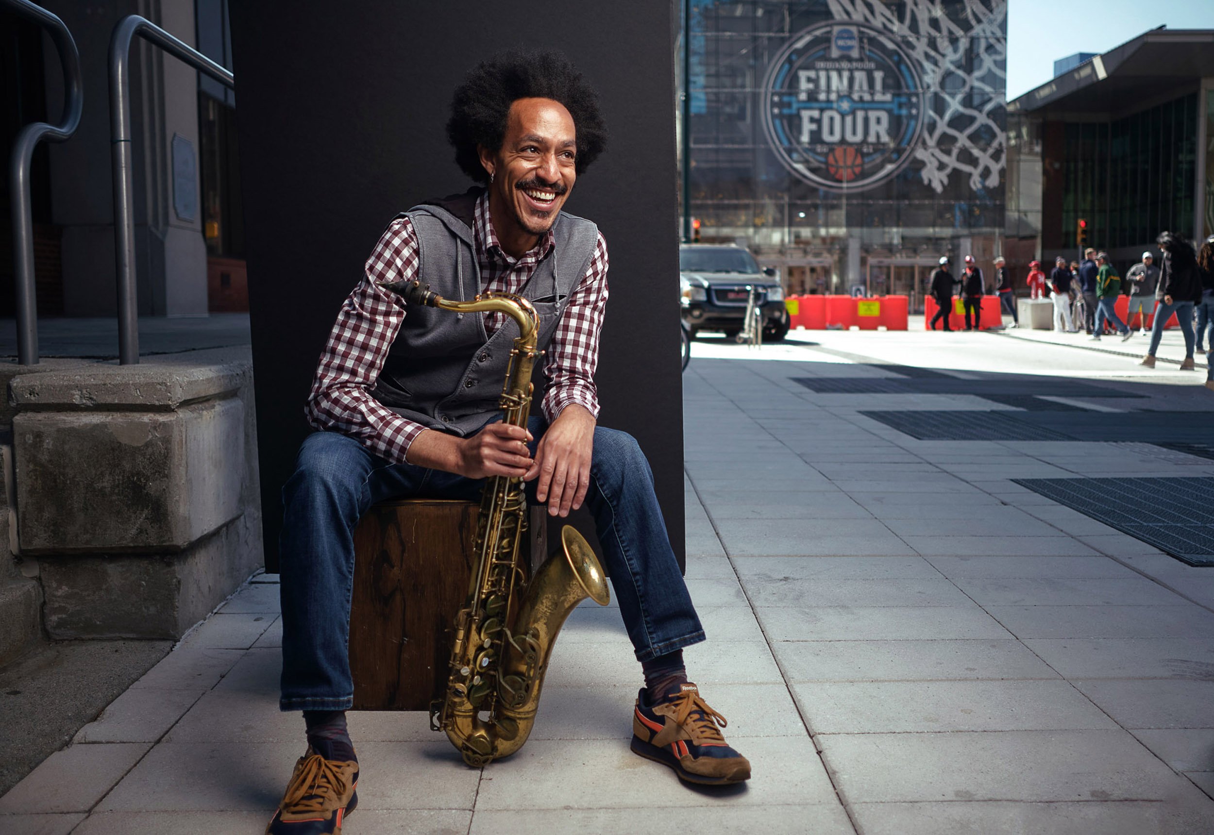 Smiling street saxophonist seated outside a downtown basketball arena with a saxophone