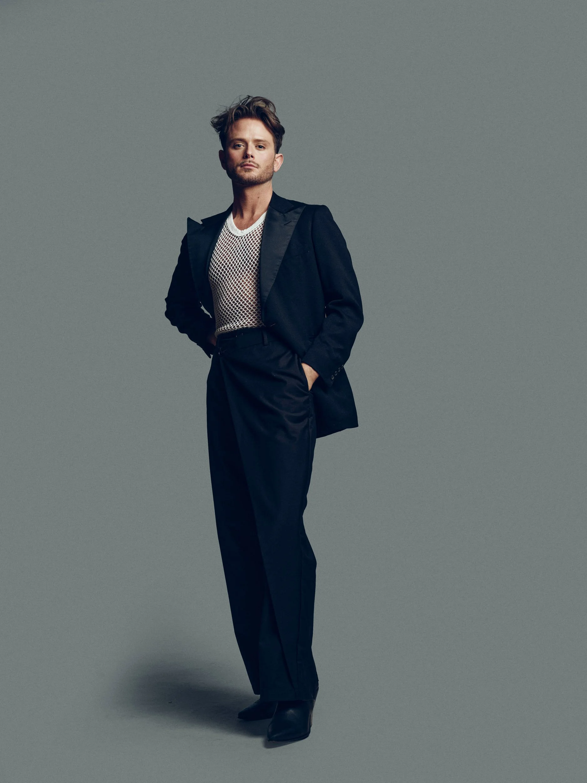 Full-length portrait of a man wearing a black suit over a white mesh shirt against a gray studio background