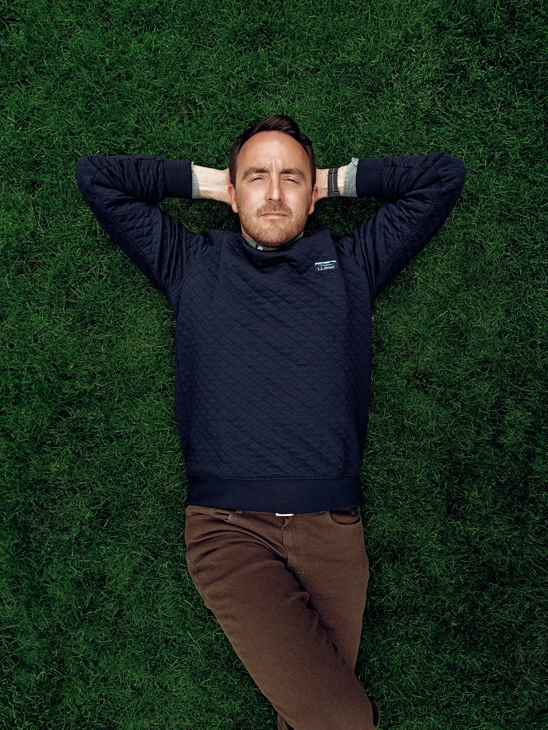 Ryan Hunley for Pattern Magazine Man relaxing on green grass with his hands behind his head, wearing a navy quilted sweater and brown pants 