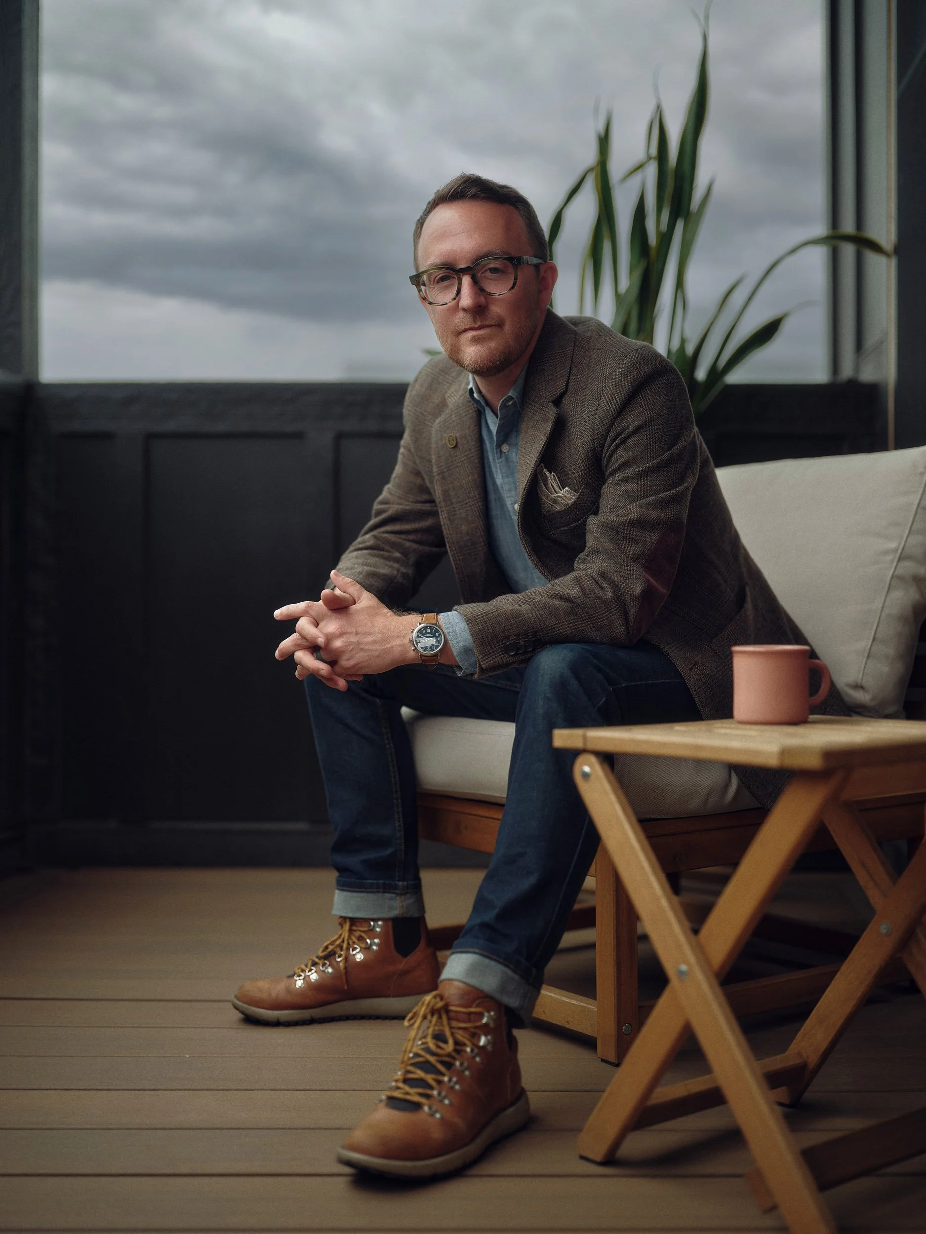 Ryan Hunley for Pattern magazine Man wearing glasses and a tweed blazer sitting in a lounge chair beside a coffee table and potted plant