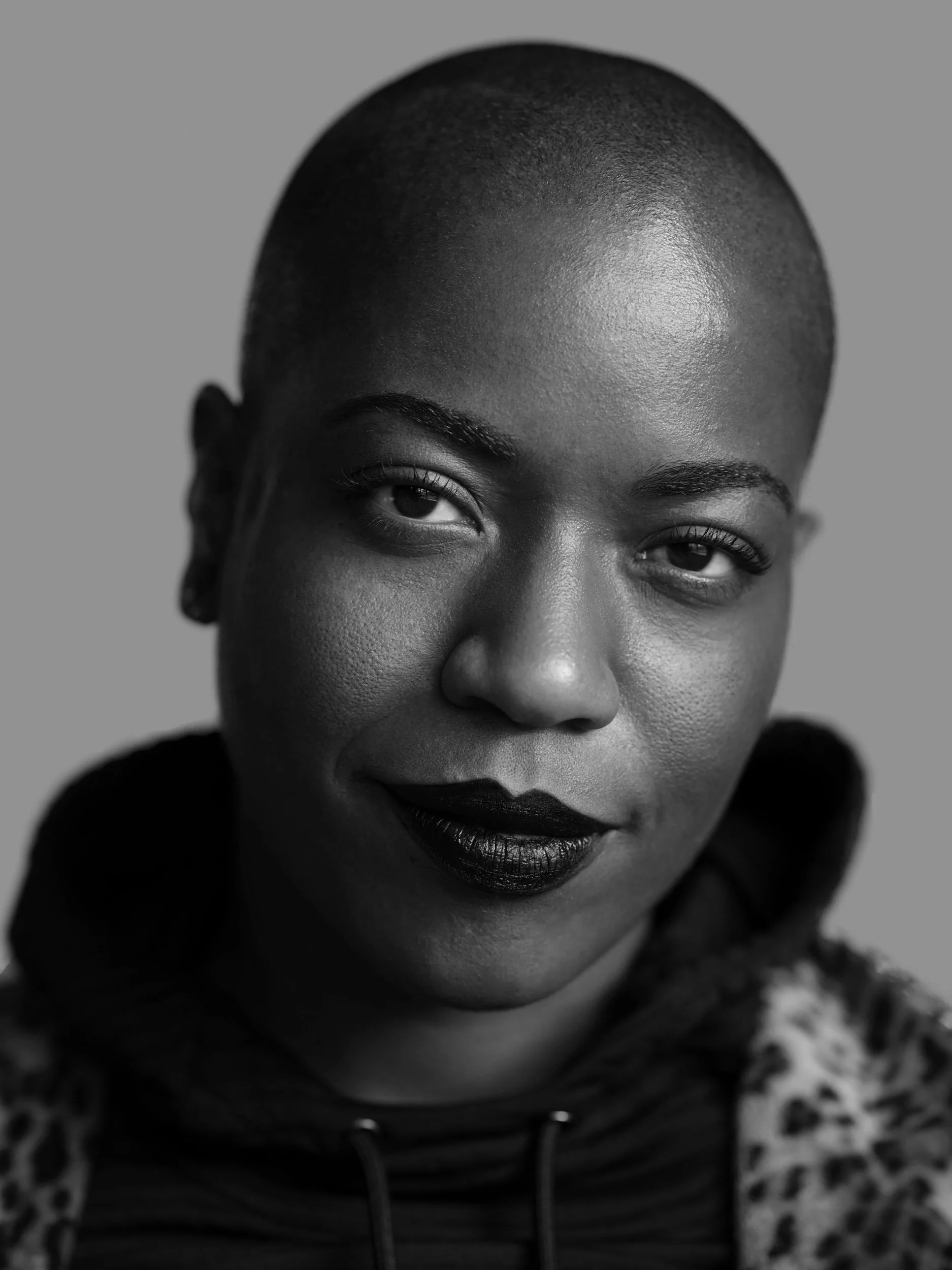 Black-and-white close-up portrait of a bald Black woman wearing dark lipstick and a patterned jacket.