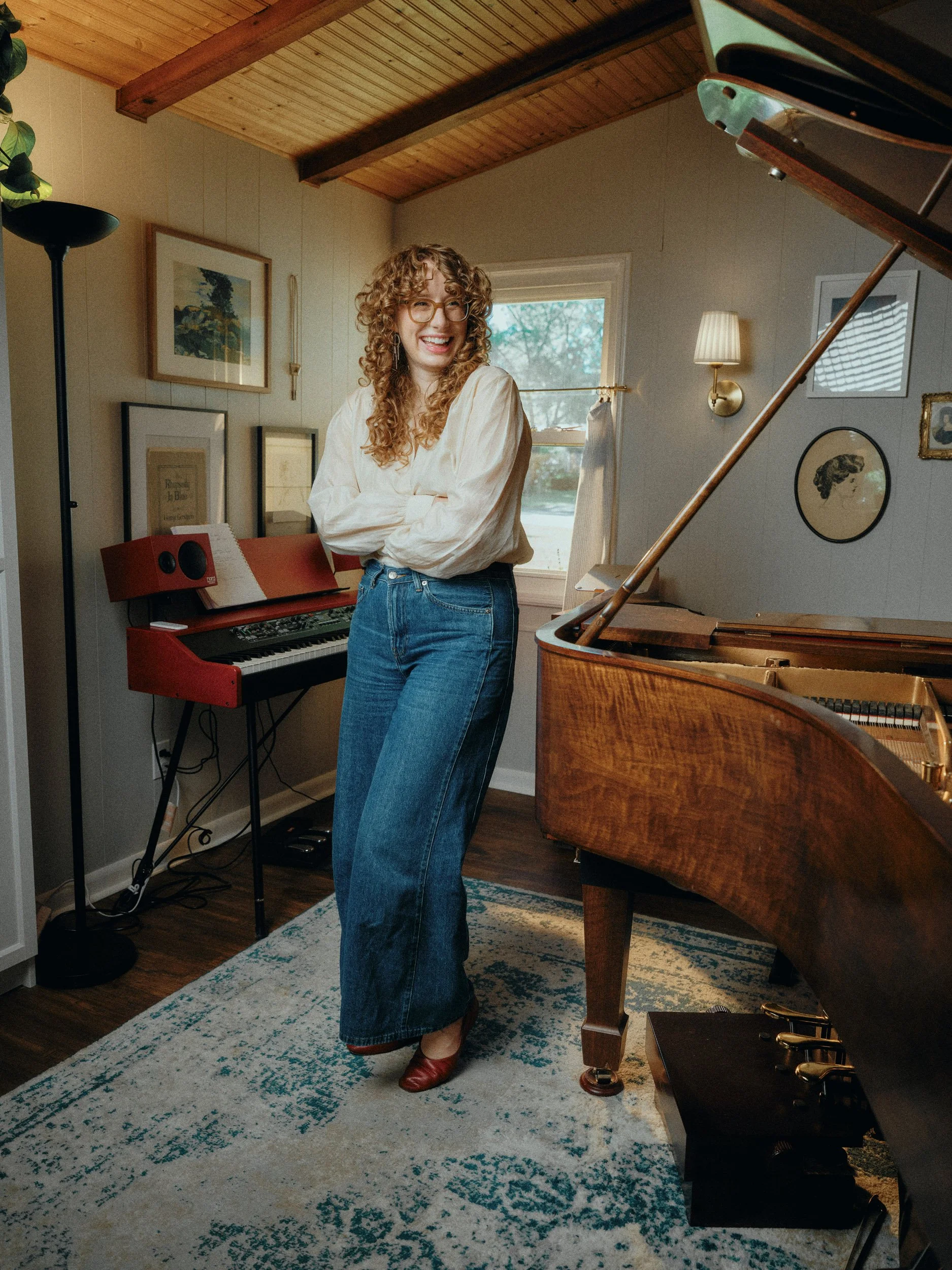Smiling woman with curly hair standing beside a grand piano and electronic keyboard in a cozy music room