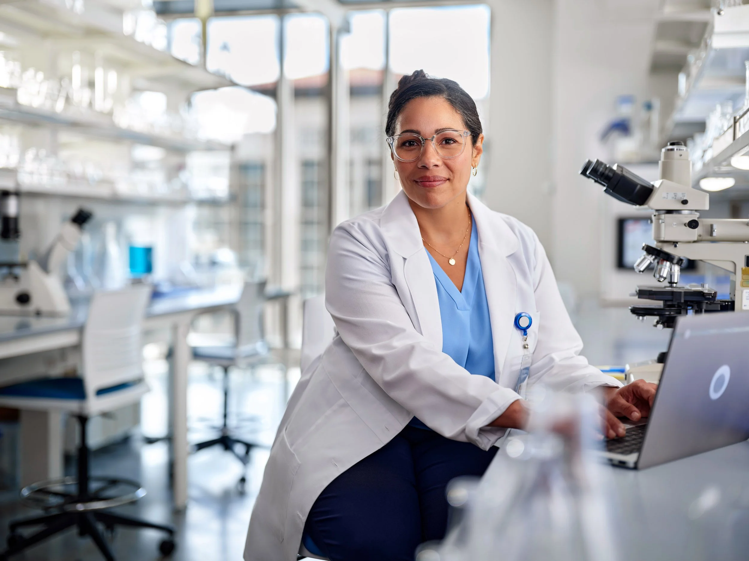 Scientist in a white lab coat working on a laptop in a laboratory with microscopes