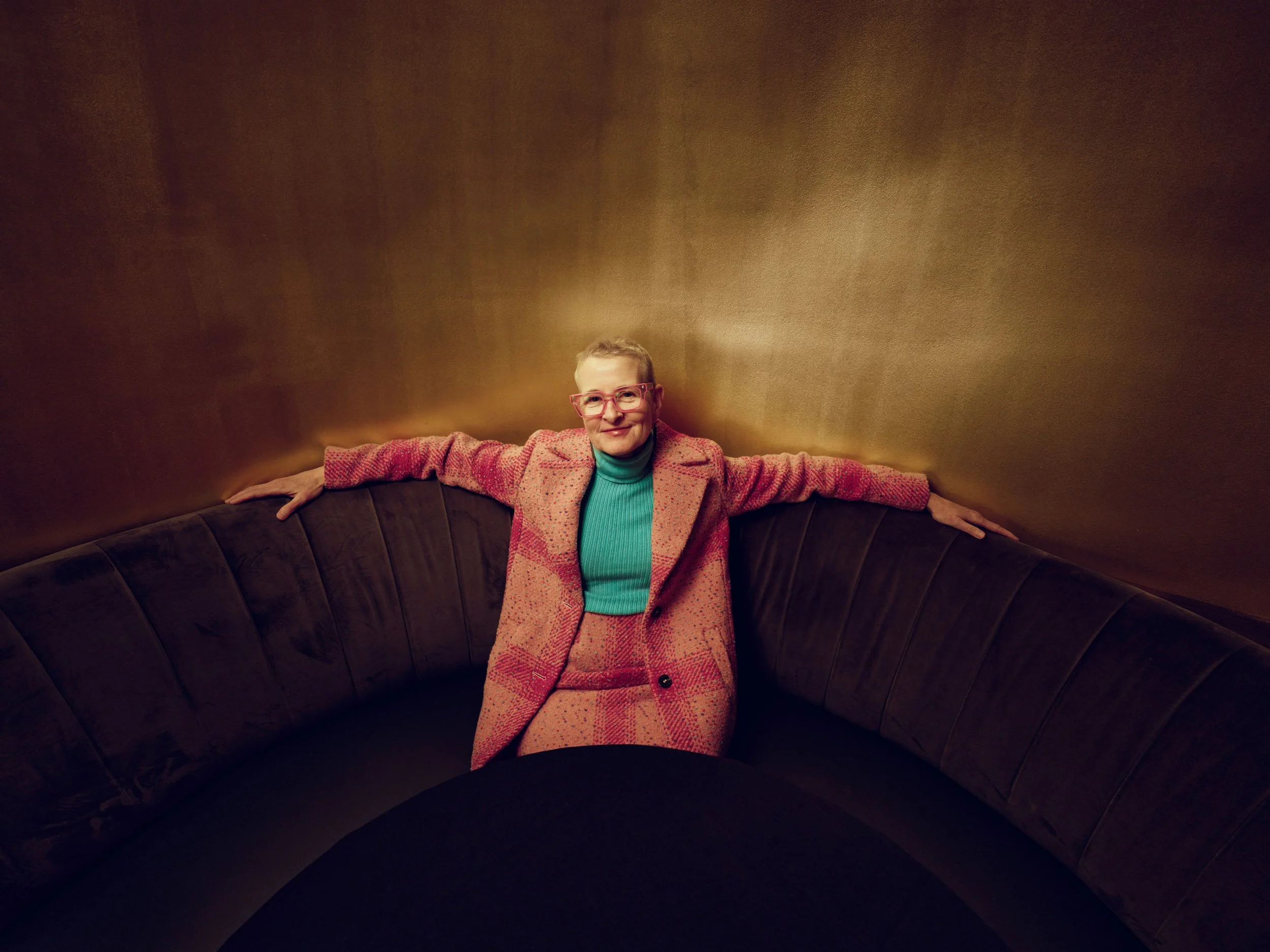 Person wearing pink glasses and a pink suit seated on a dark curved sofa against a warm brown wall