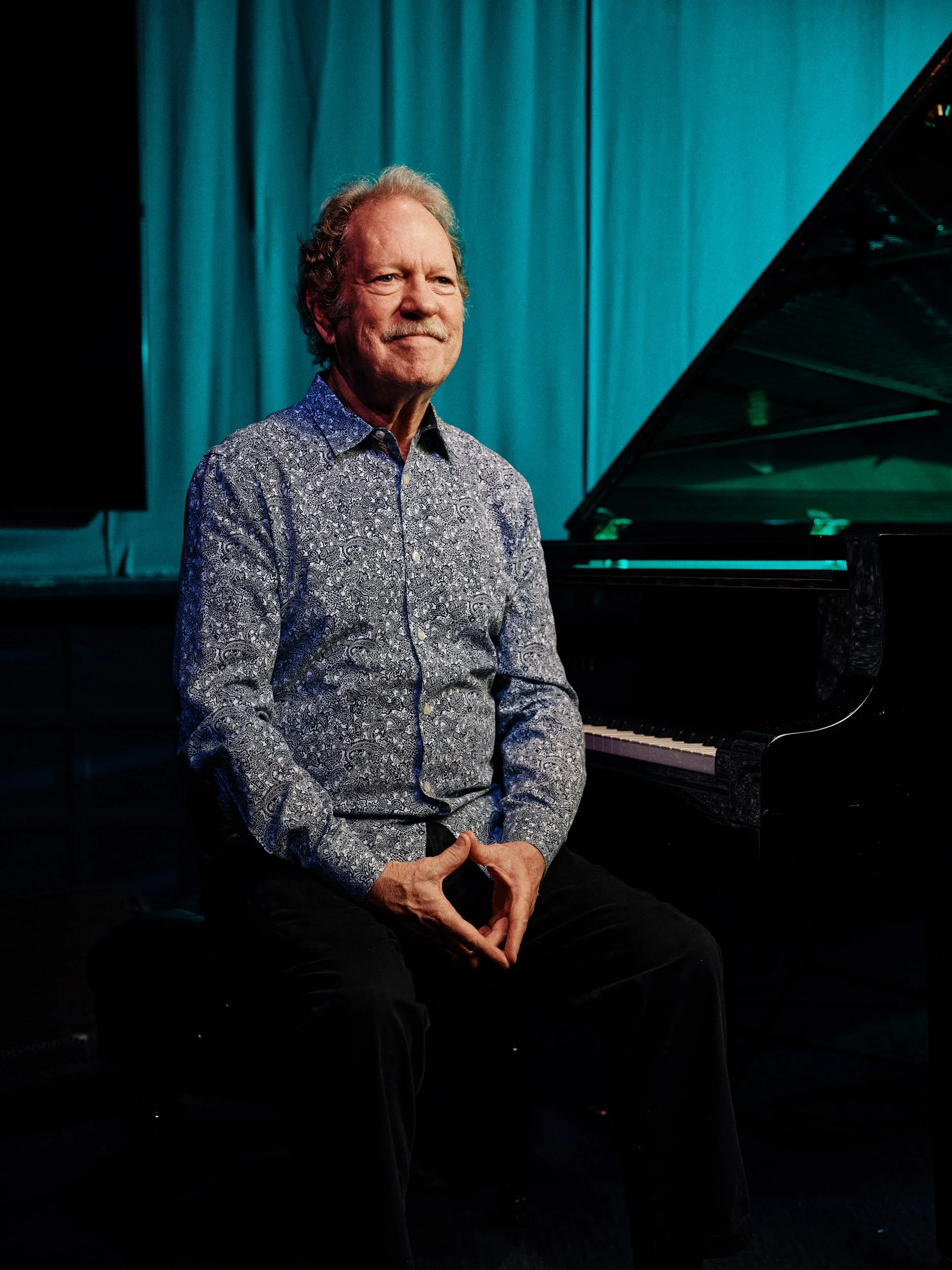 Man in a patterned shirt seated beside a grand piano on a stage with teal curtains Jazz Player