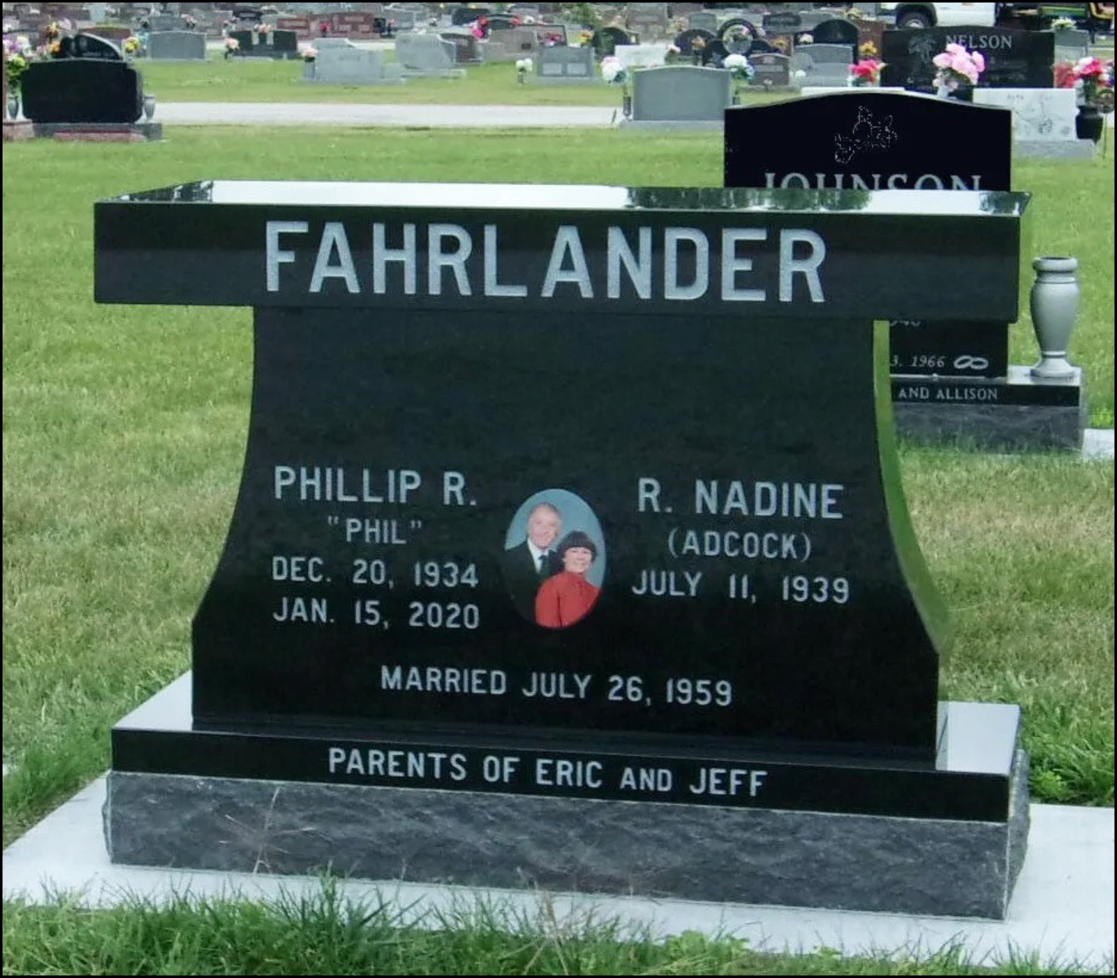 Black grave marker with white and gray lettering for Phillip R. Fahrlander and R. Nadine Fahrlander, includes their birth and death dates, a photo of the couple, and mentions their marriage date, with surrounding graves and grass.