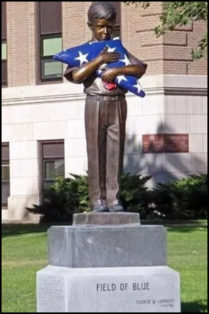 Statue of a young boy holding an American flag, standing on a stone pedestal outside a brick building.