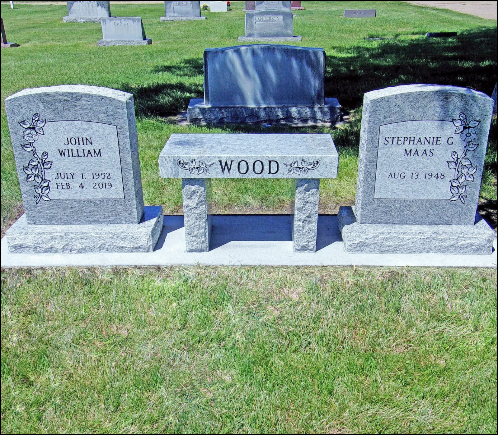 Gravestone monument in a cemetery with the surname Wood engraved on a central bench and two headstones on either side for John William Wood and Stephanie G. Maas, each decorated with floral patterns.