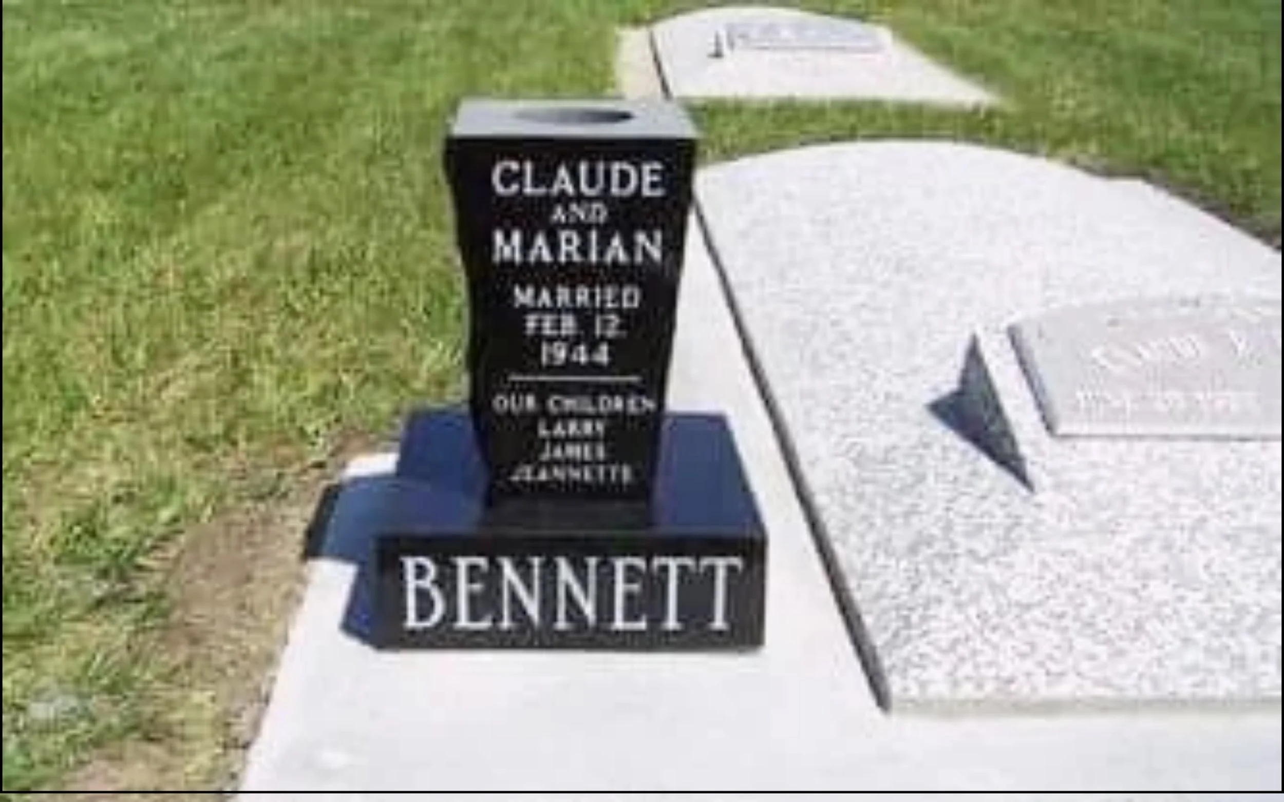 Pick up truck with black tombstone-shaped marker for Claude and Marian Bennett, with dates February 12, 1944, on a sidewalk next to grass.