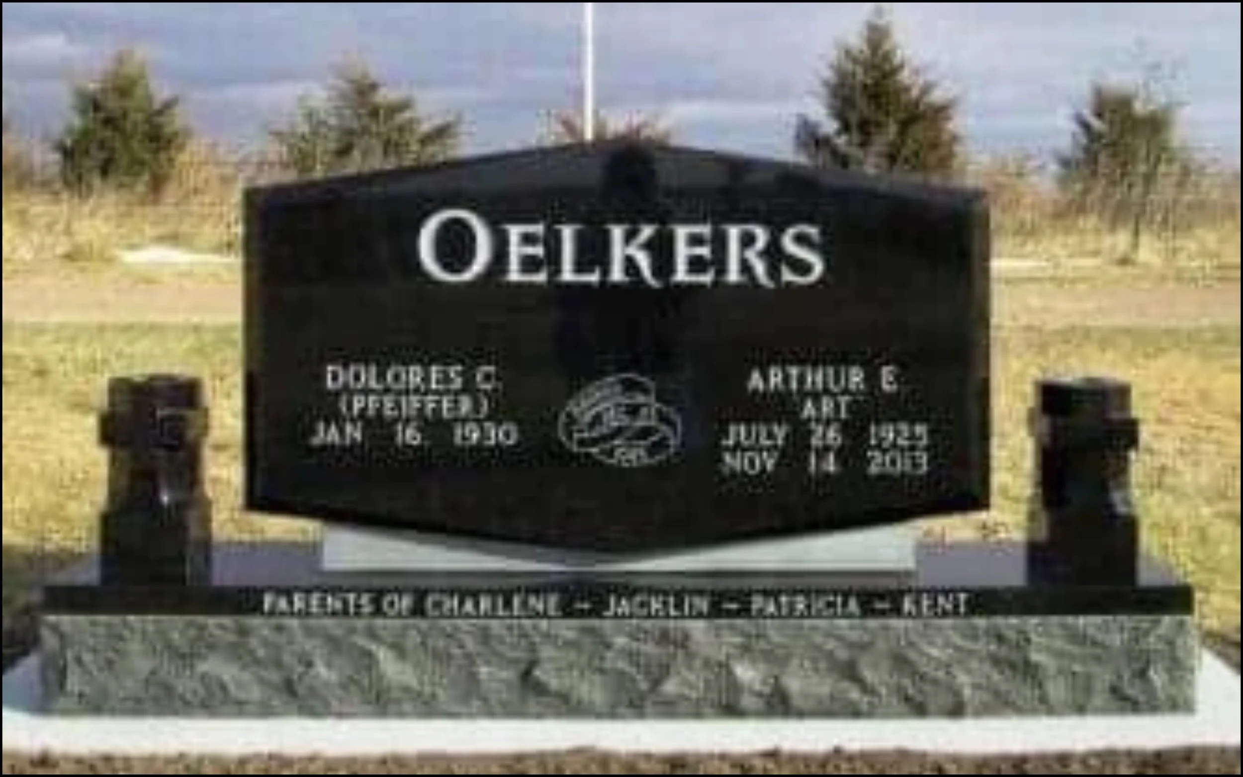 Black memorial plaque with white inscriptions honoring Oelkers. Names listed include Dolores C. and Jacklin P. with dates, along with Arthur G. and other names, with a background of trees and cloudy sky.