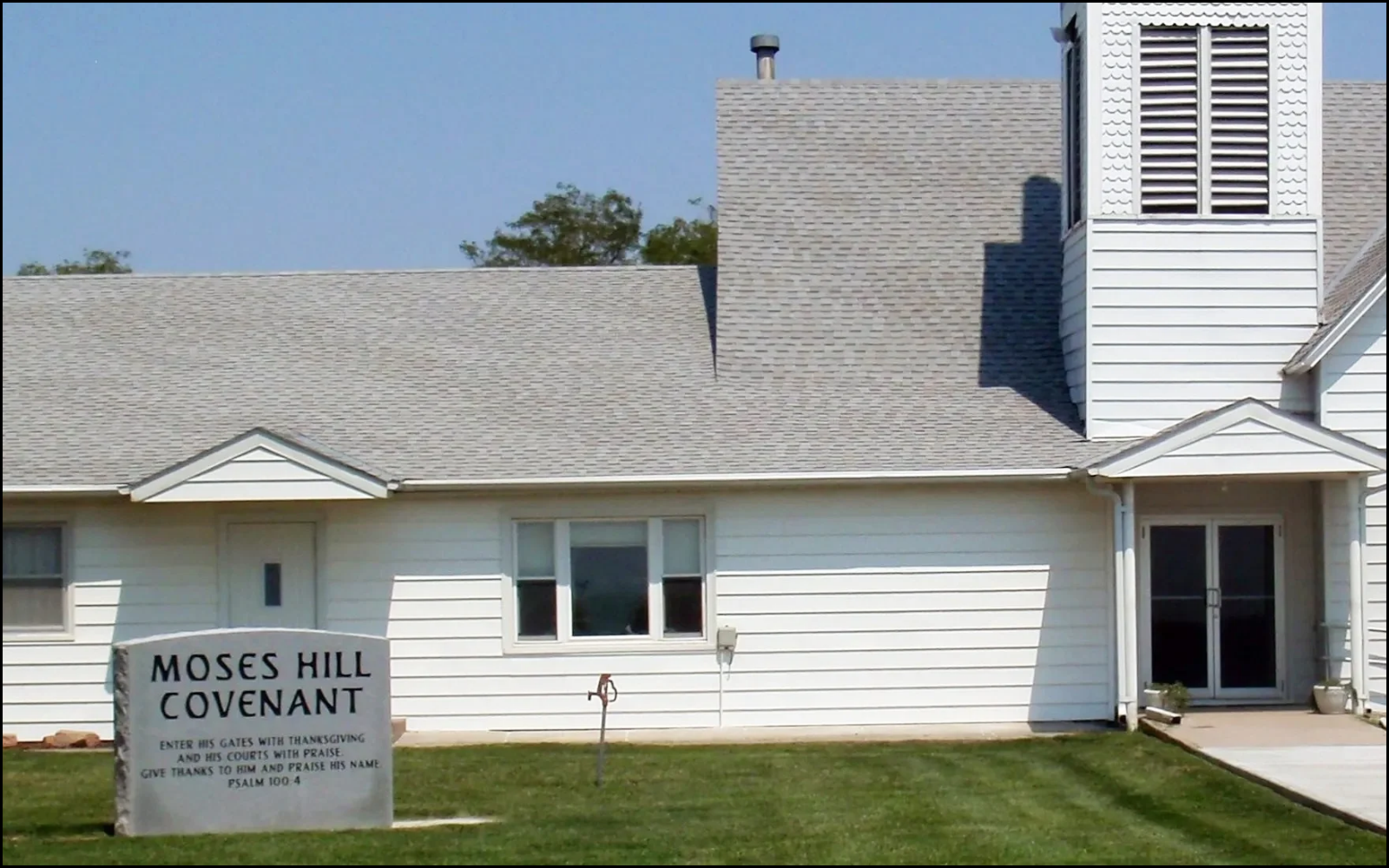 A white church building with a gray shingled roof, includes a sign reading 'Moses Hill Covenant' with a religious message, and a grassy lawn in front.